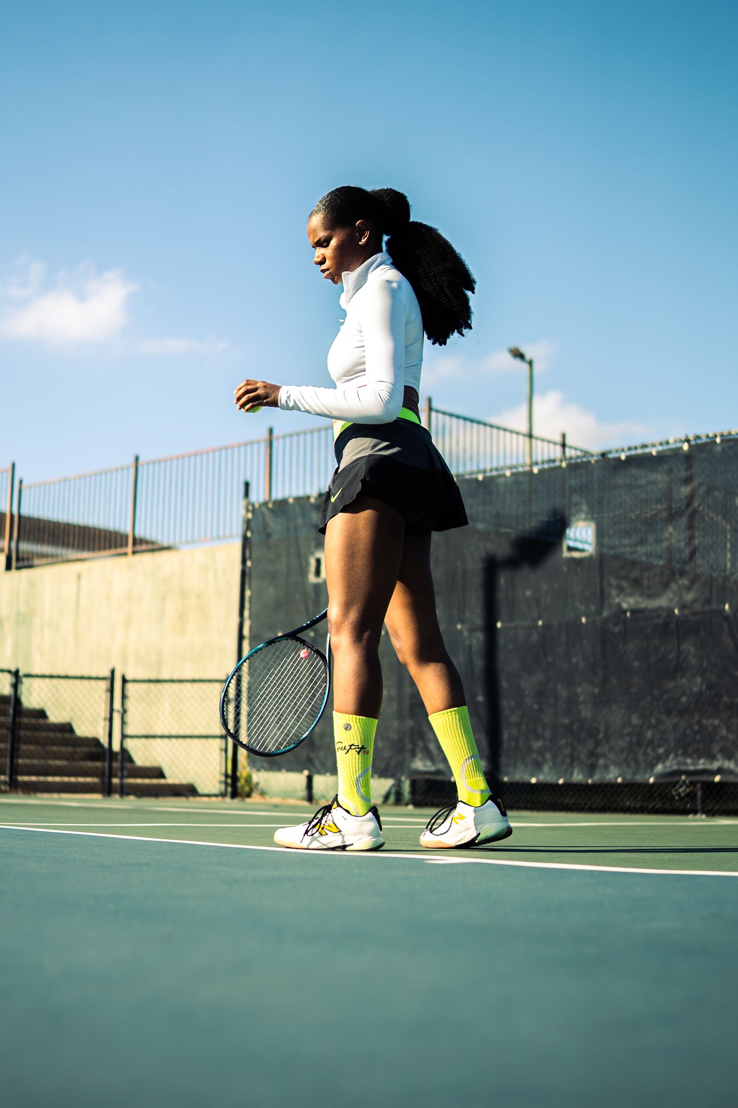 A young Black woman in tennis attire with neon green socks holds a racket on an outdoor court under a blue sky.