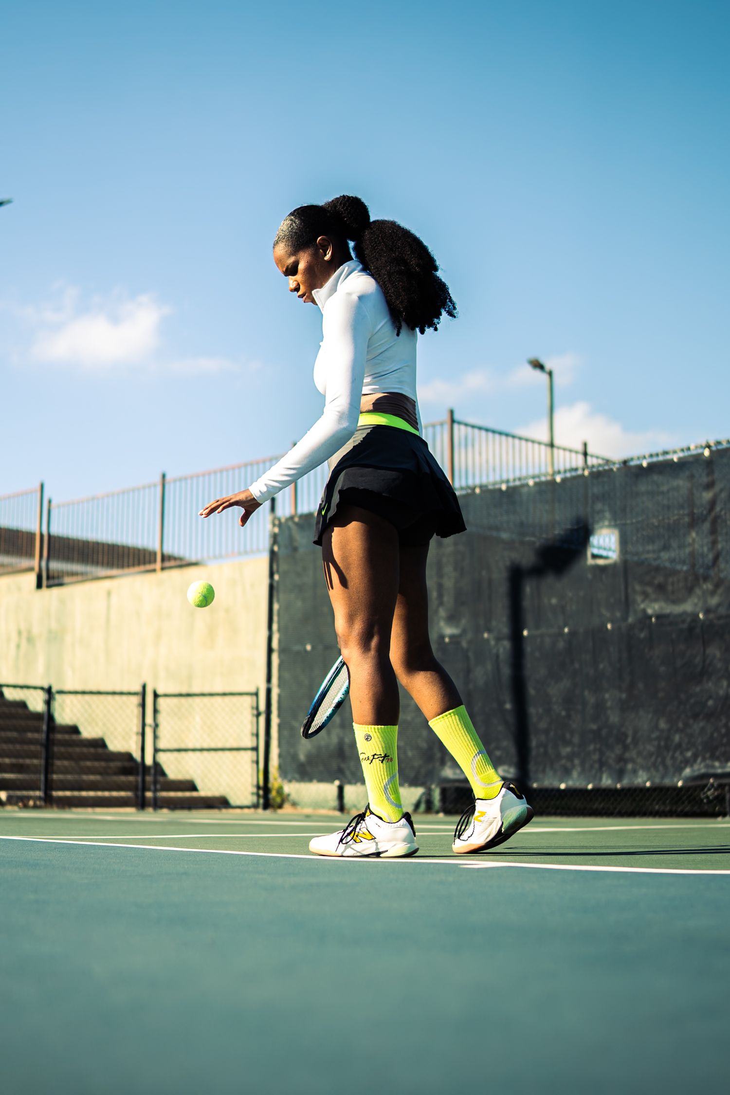 Female tennis player in white top and neon green socks bouncing ball on blue outdoor court.