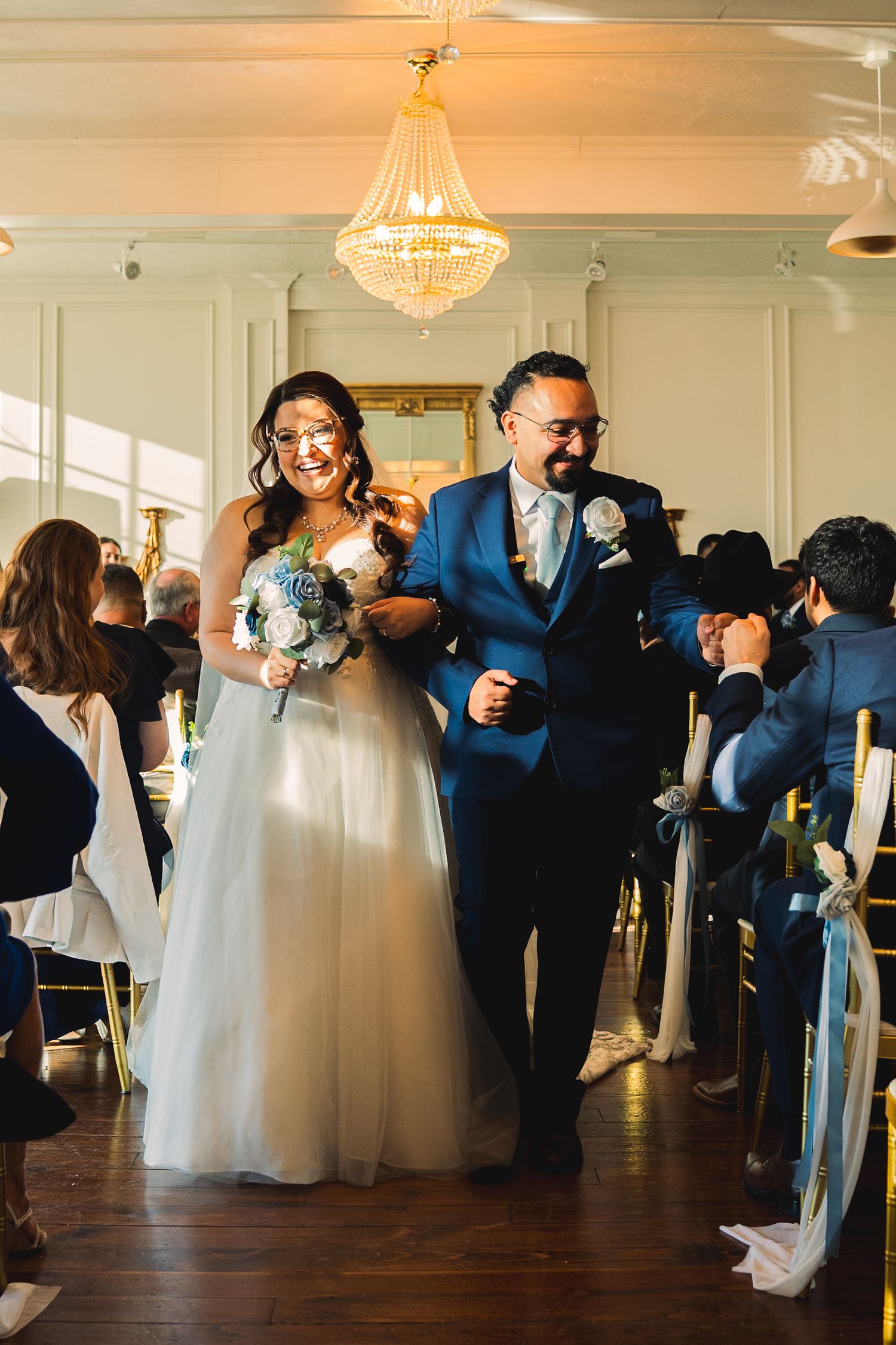 Smiling bride and groom walk down the aisle in an elegant chandelier-lit indoor ceremony.