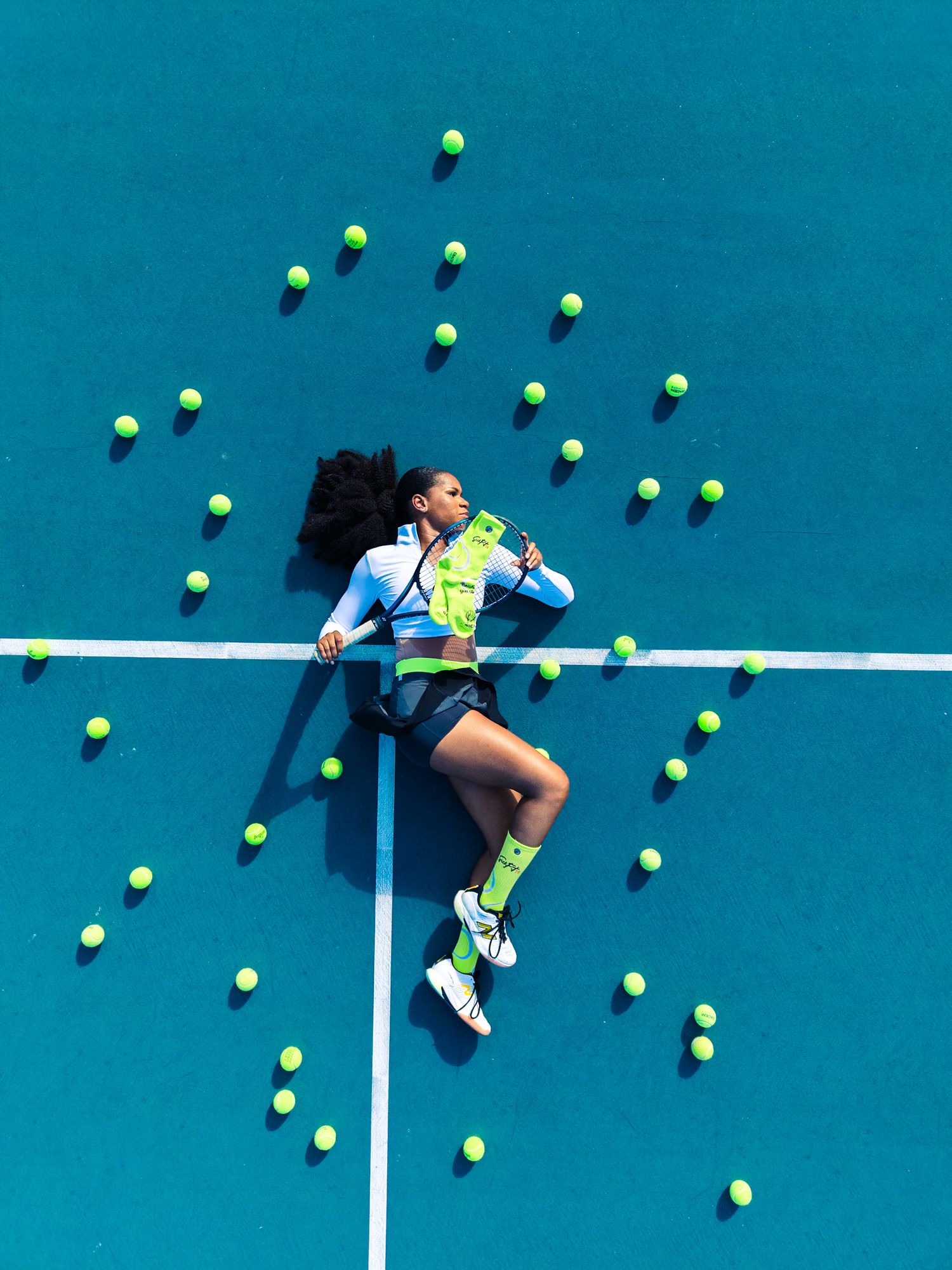 Aerial view of a female tennis player lying on a blue court surrounded by scattered neon yellow tennis balls.