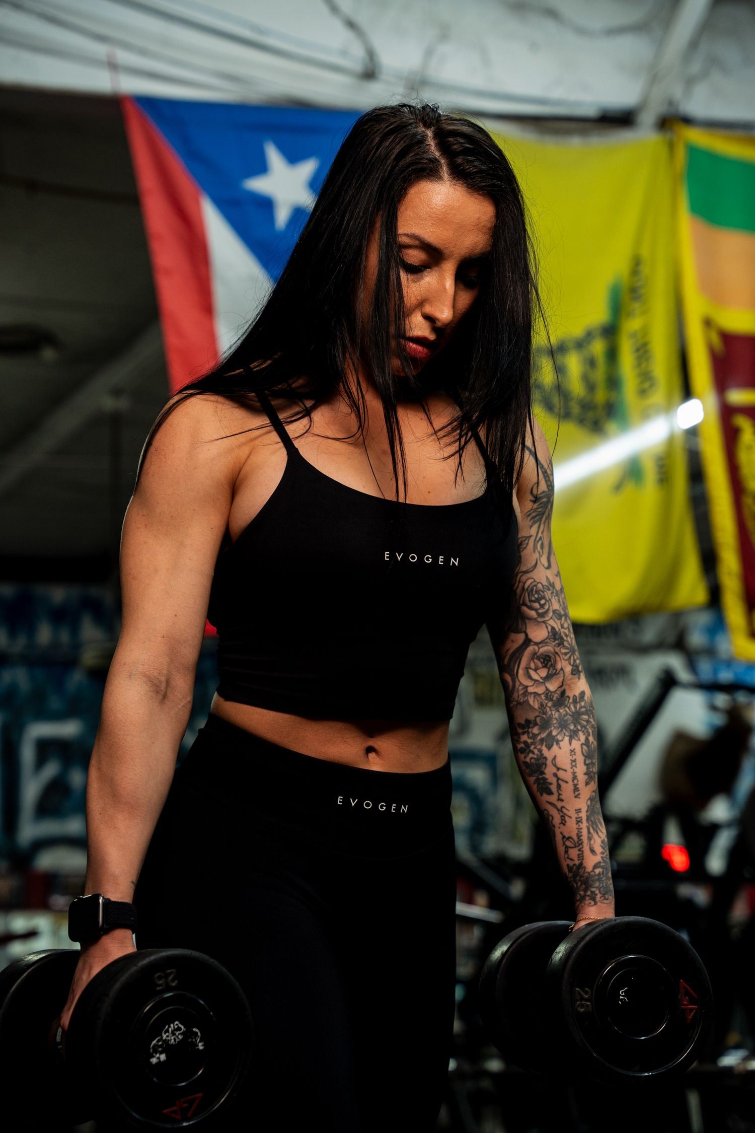 A tattooed woman in black Evogen workout set lifts dumbbells in a gym with colorful flags.