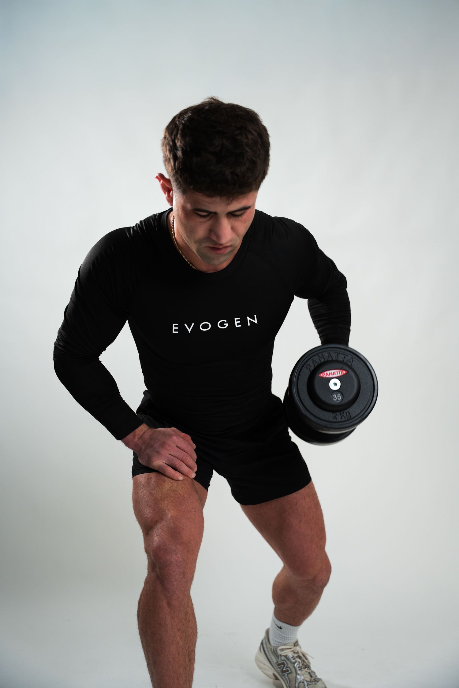Muscular man in black Evogen long sleeve shirt curling dumbbell against white studio background.