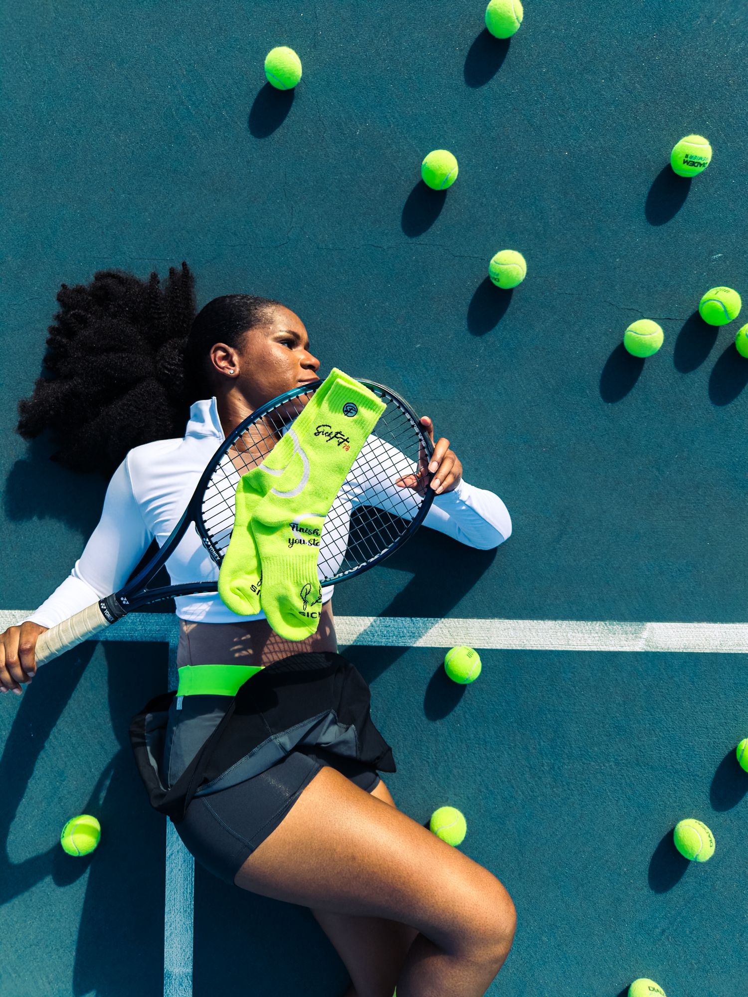 Woman in tennis outfit lying on blue court surrounded by tennis balls, holding racket and neon green socks.