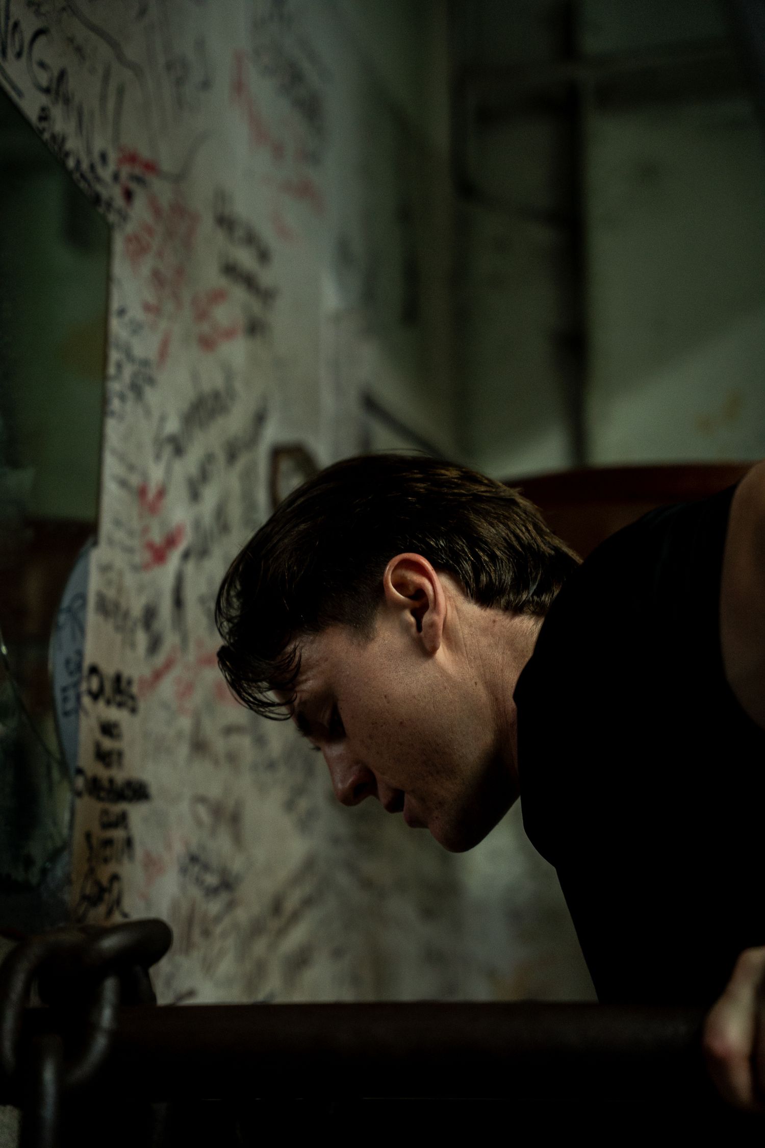 A young man with dark hair bows his head in a dimly lit gym with graffiti-covered walls.