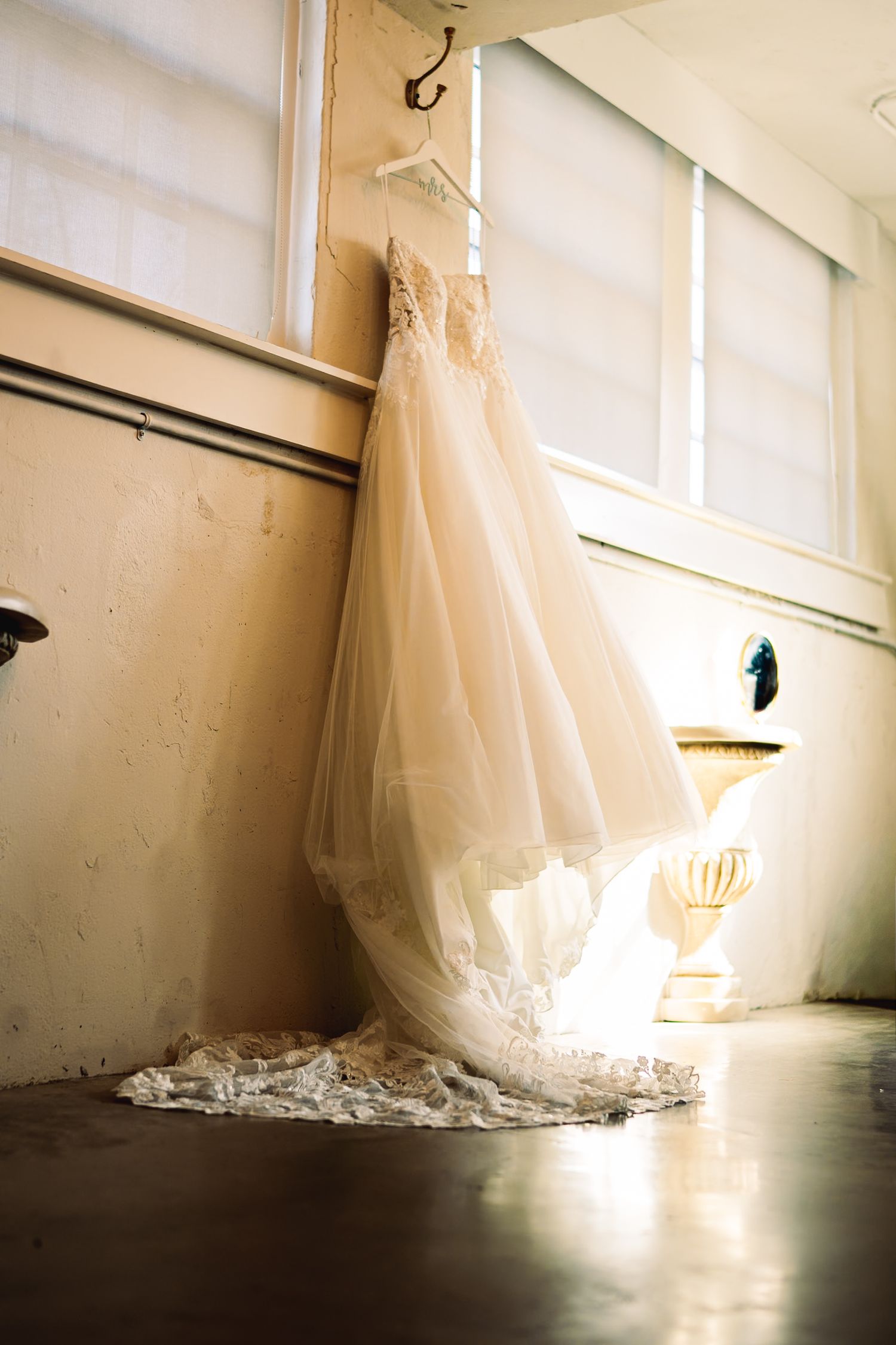 Elegant lace wedding gown hanging by window in soft natural light.