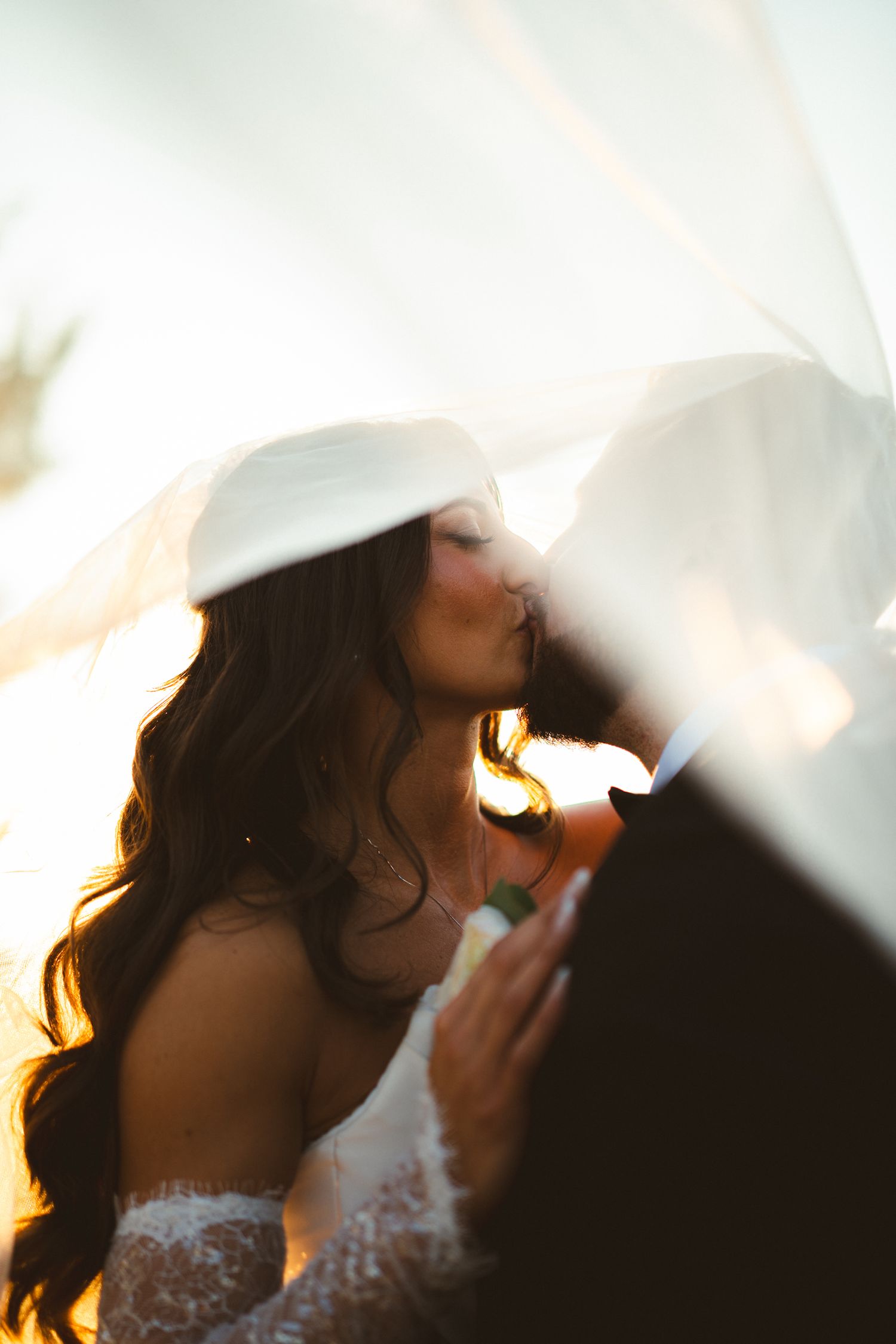 Bride in lace dress and white hat kissing groom in golden sunset backlit by flowing veil.