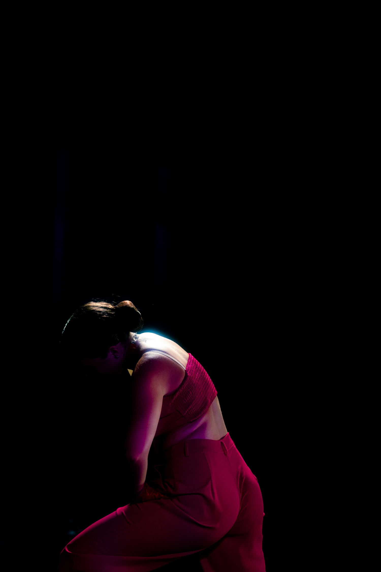 A dancer in a red outfit bends forward dramatically under a beam of light against a dark black background.