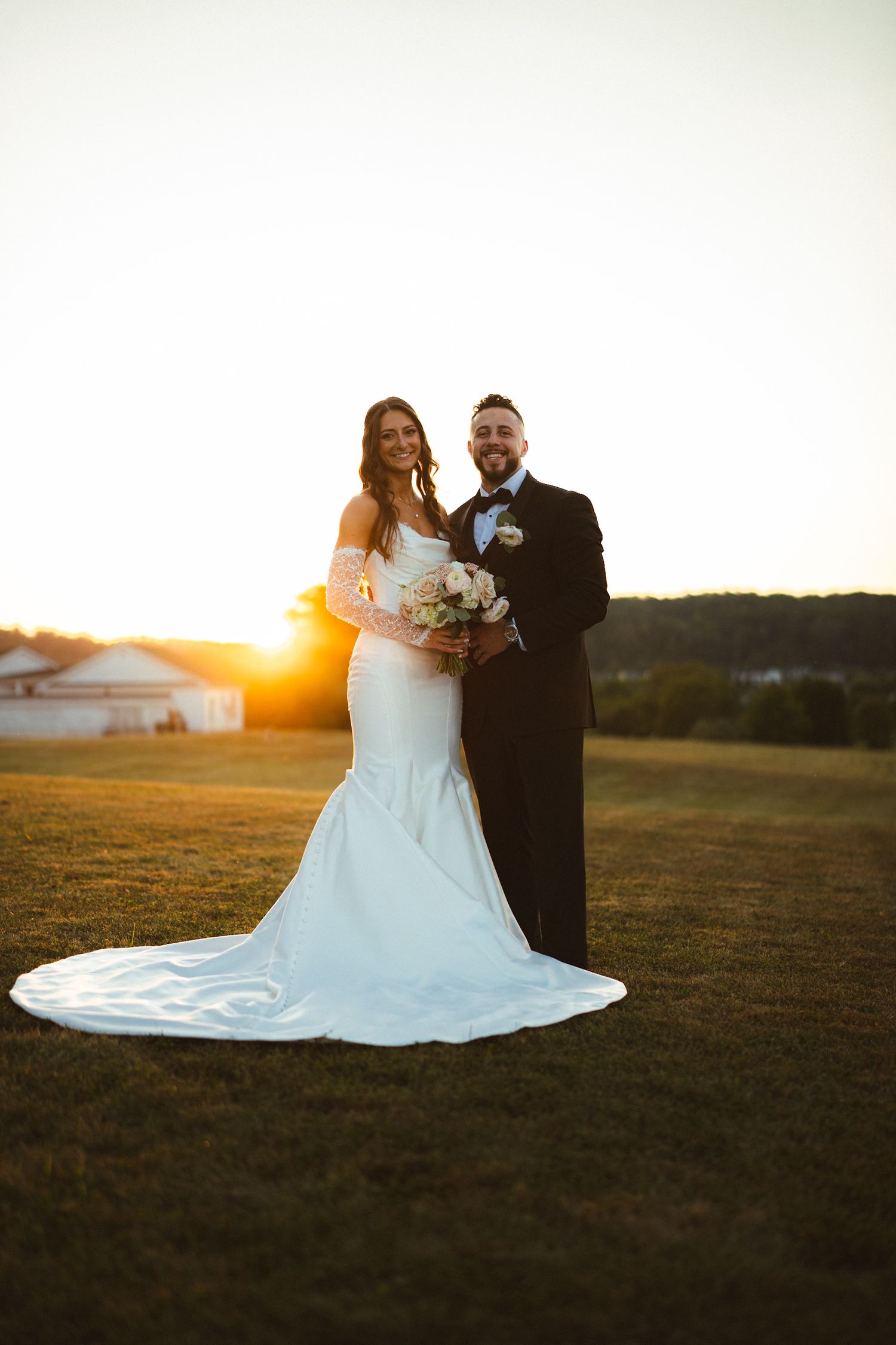 Bride and groom pose together at sunset in a field, bride holding a white bouquet.
