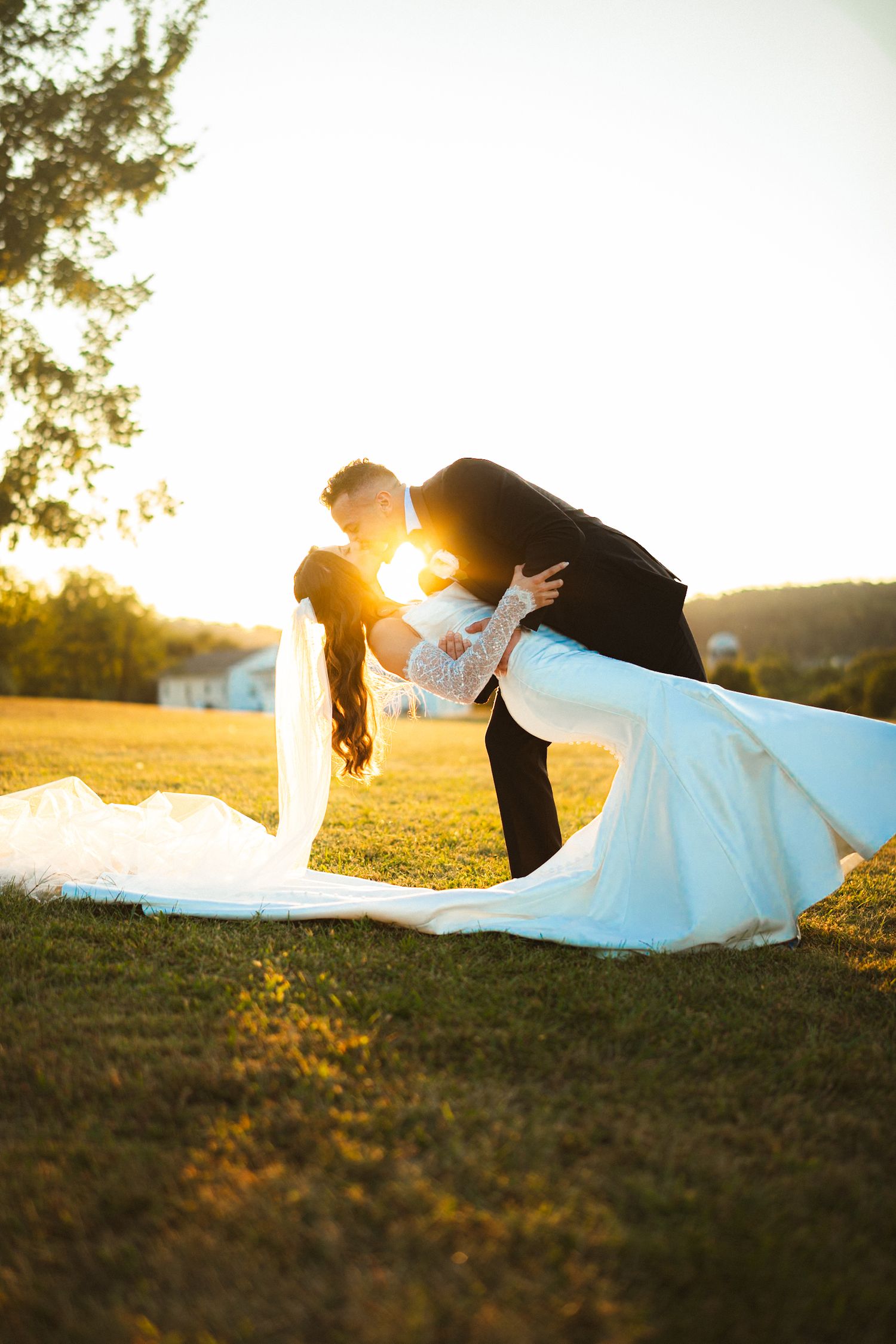 Groom dips bride in romantic kiss on green field at golden hour, wedding dress and veil trailing gracefully.