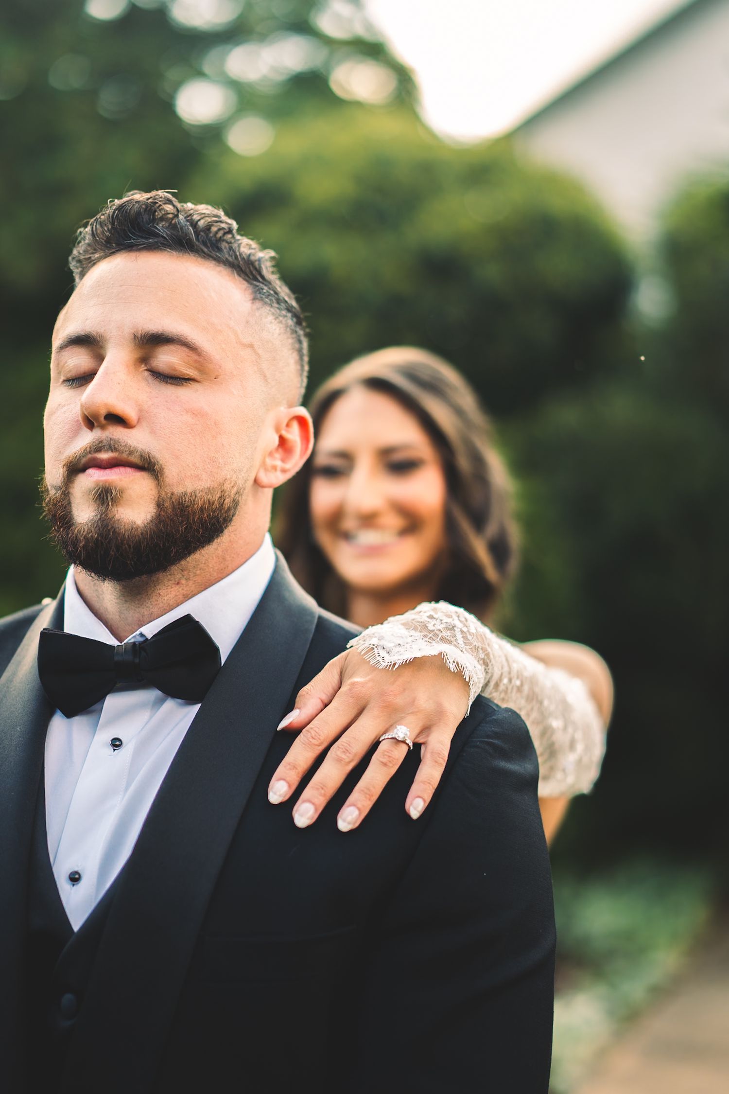 Smiling bride in lace gown places hand on grooms shoulder as he poses in black tuxedo with bow tie.