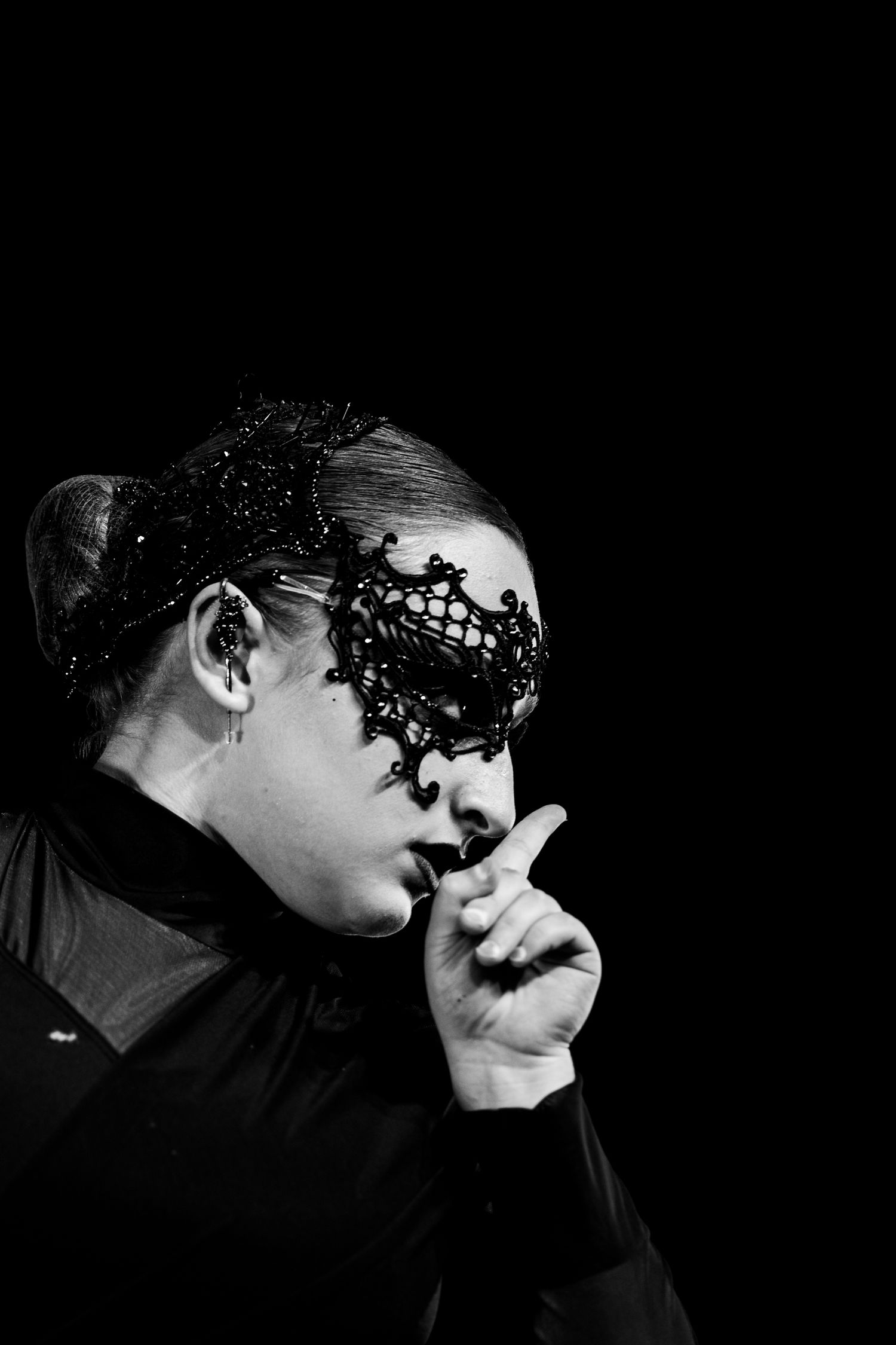 Black and white photo of a woman wearing an ornate black lace masquerade mask, holding finger to lips in silence gesture.