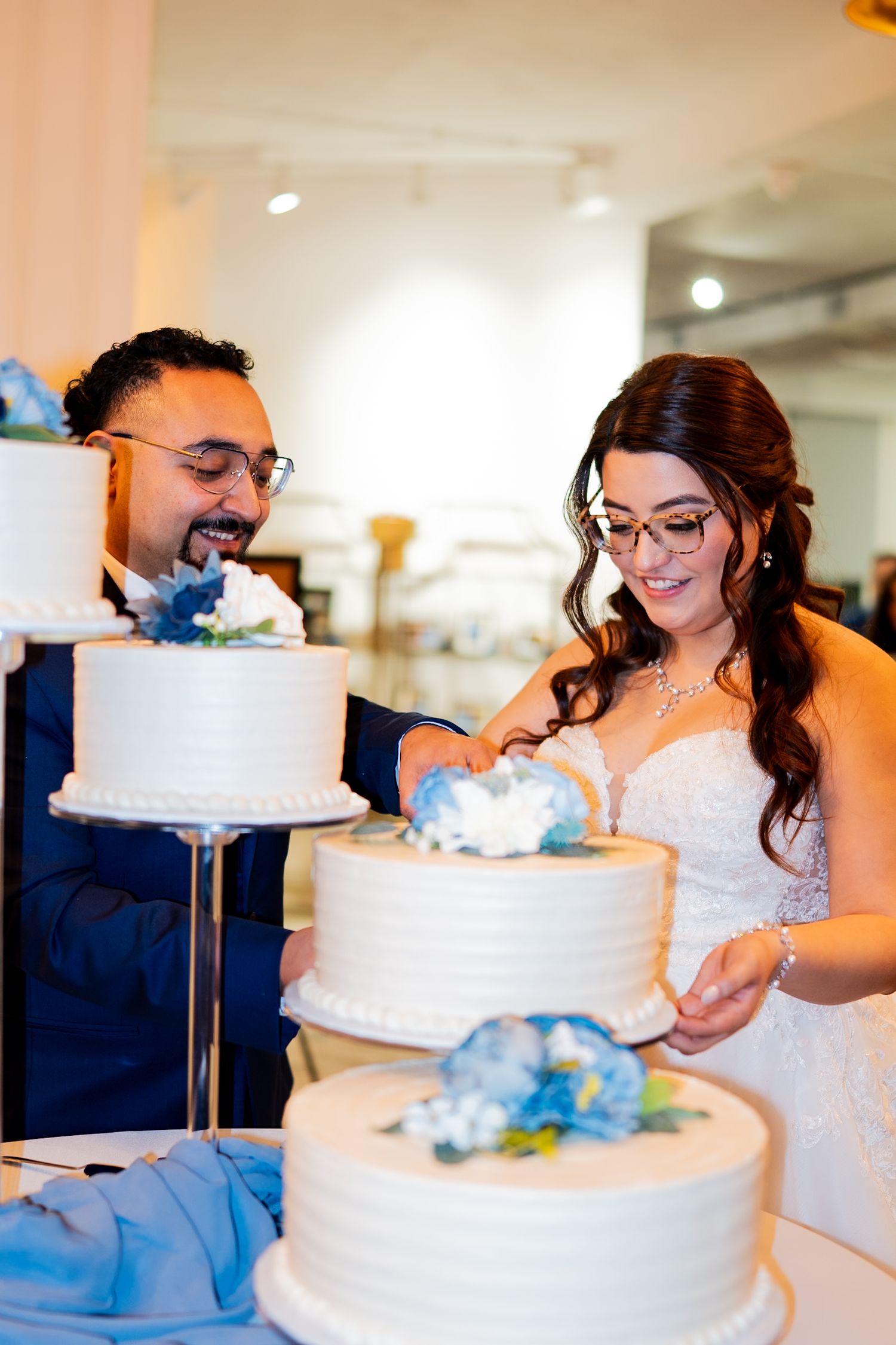 Bride and groom with glasses cut a tiered white wedding cake decorated with blue flowers.
