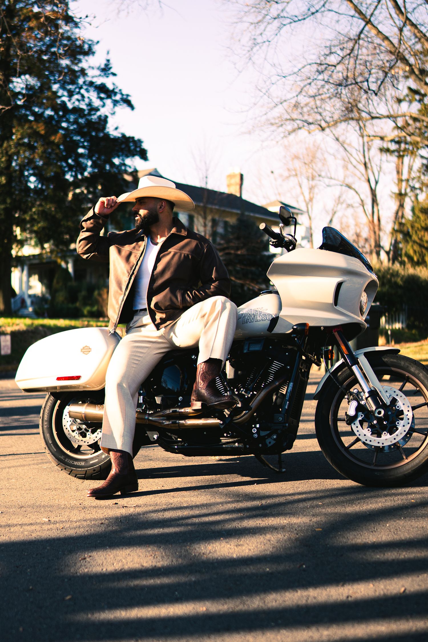 Man in cowboy hat and brown jacket posing on a white Harley-Davidson motorcycle on a sunny suburban street.