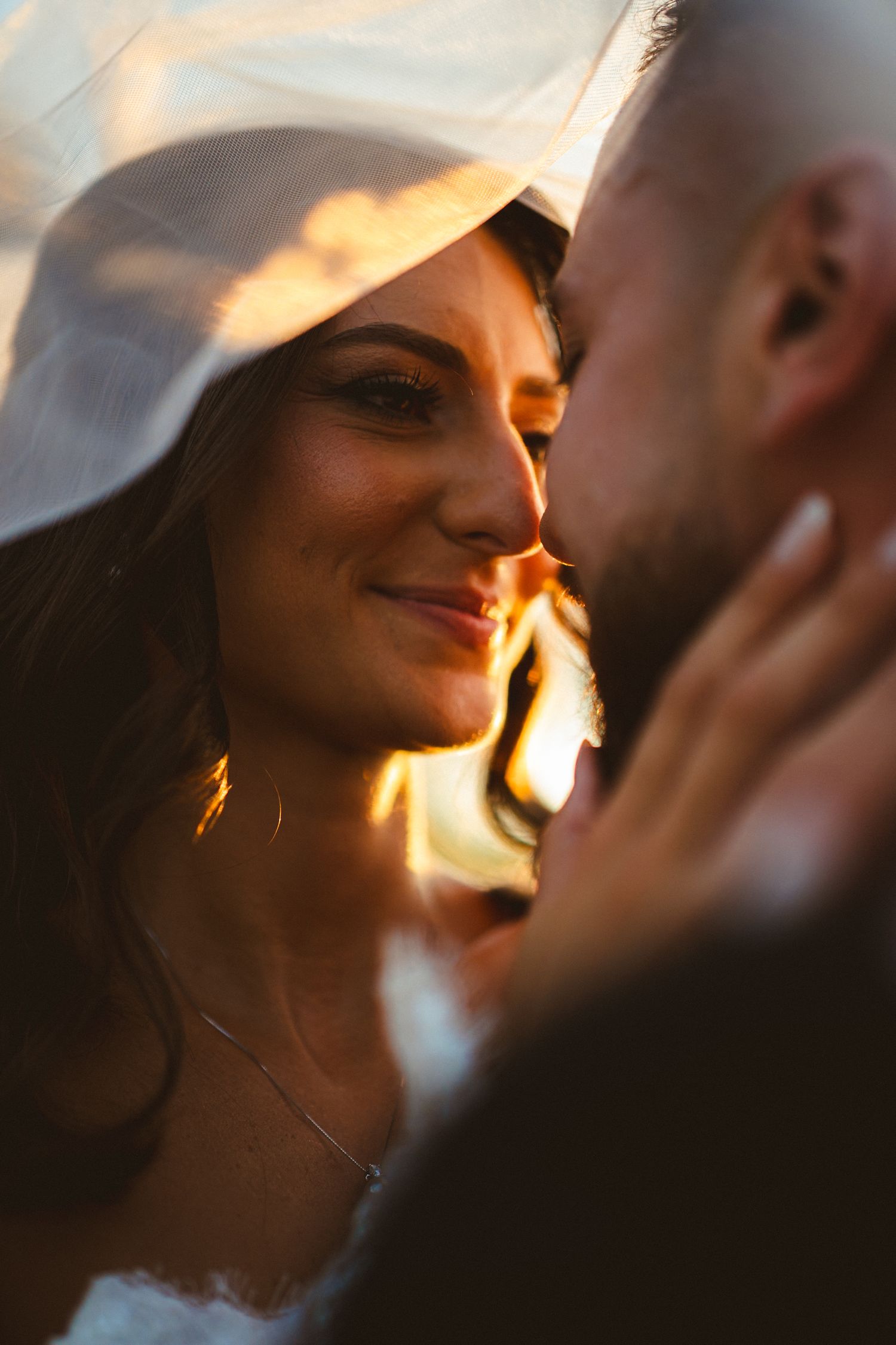 Beautiful bride smiles tenderly at groom under flowing veil in warm golden sunset light on wedding day.