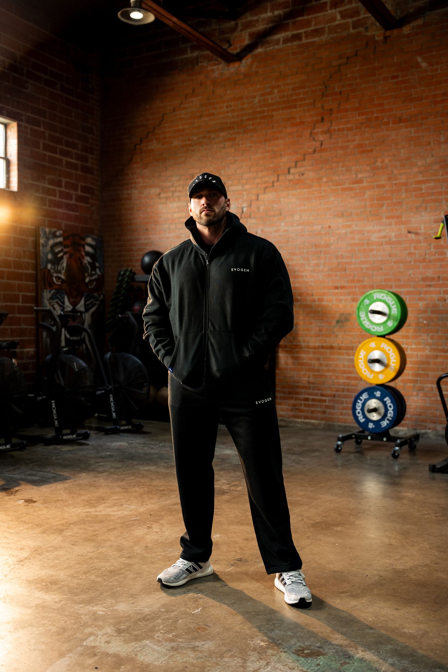 Man in black Evogen hoodie and cap standing in brick-walled gym.