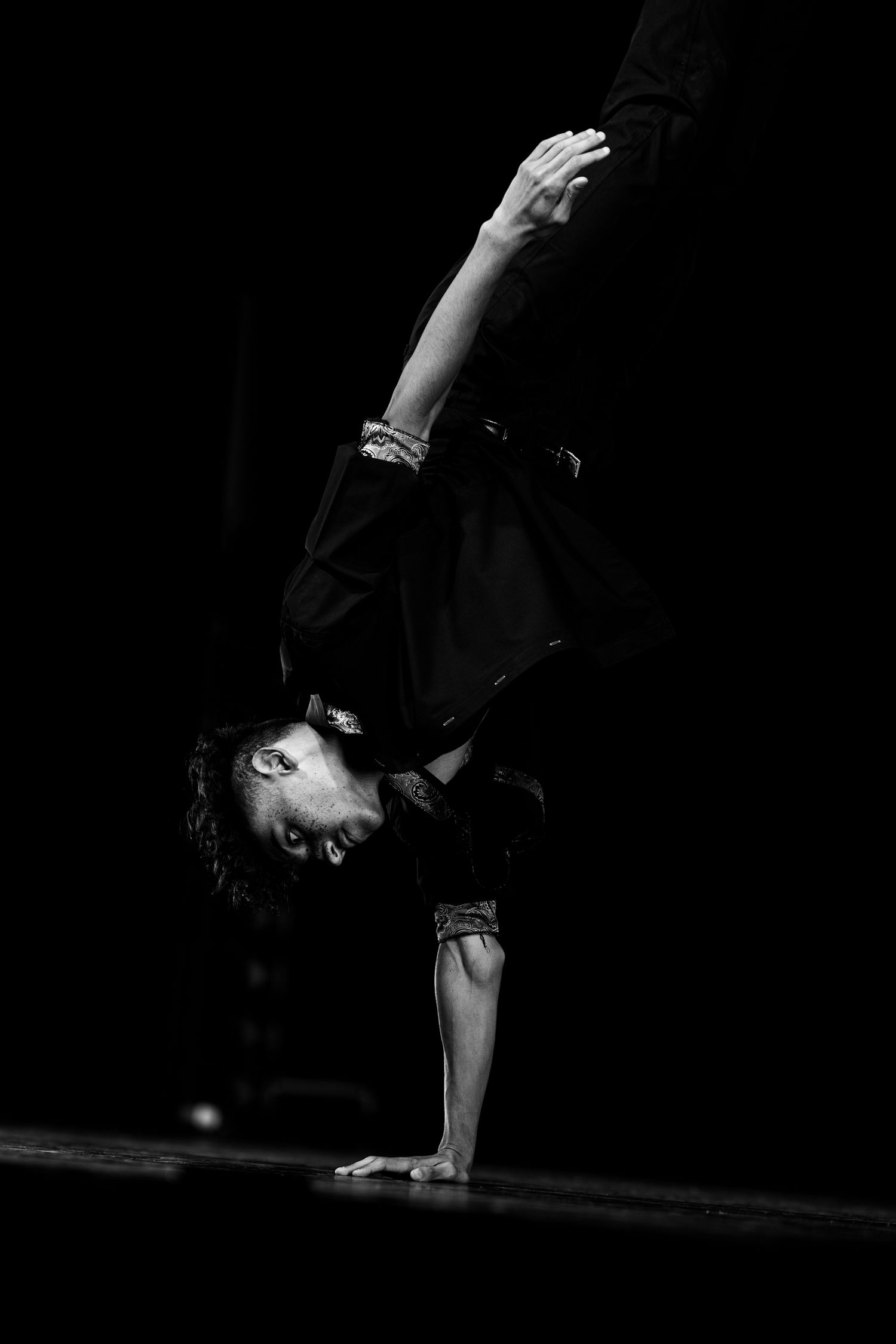 Black and white photo of a breakdancer performing a one-handed handstand on a dark stage.