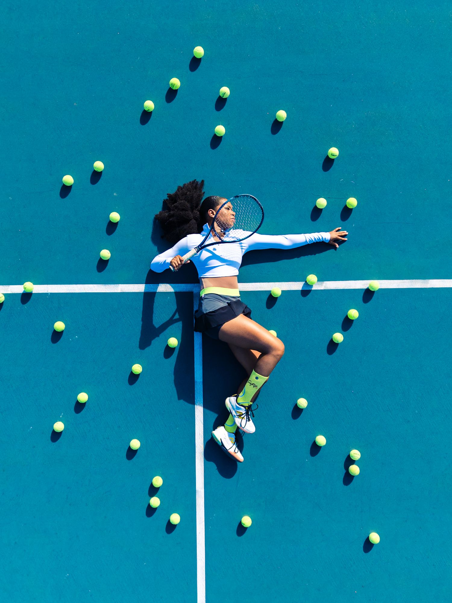 Aerial view of a female tennis player lying on a blue court surrounded by scattered yellow tennis balls.