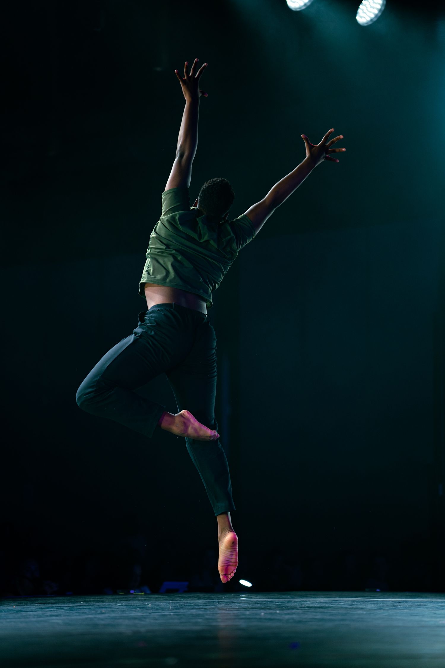 A dancer leaps gracefully on a dark stage, arms raised, wearing a green top and black leggings under dramatic spotlights.