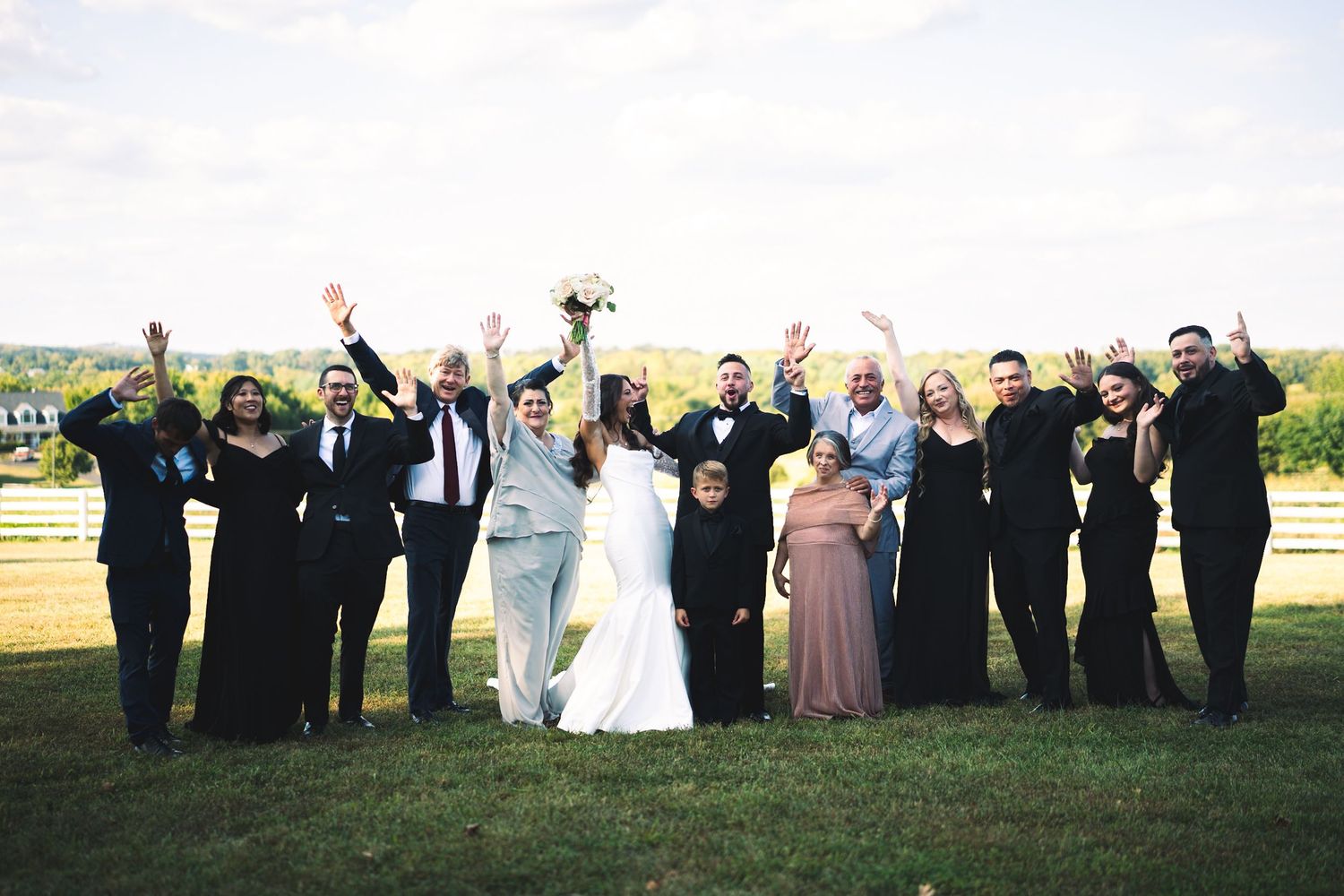 Joyful wedding party celebrates on green hillside with raised arms, dressed in black and white formal attire.