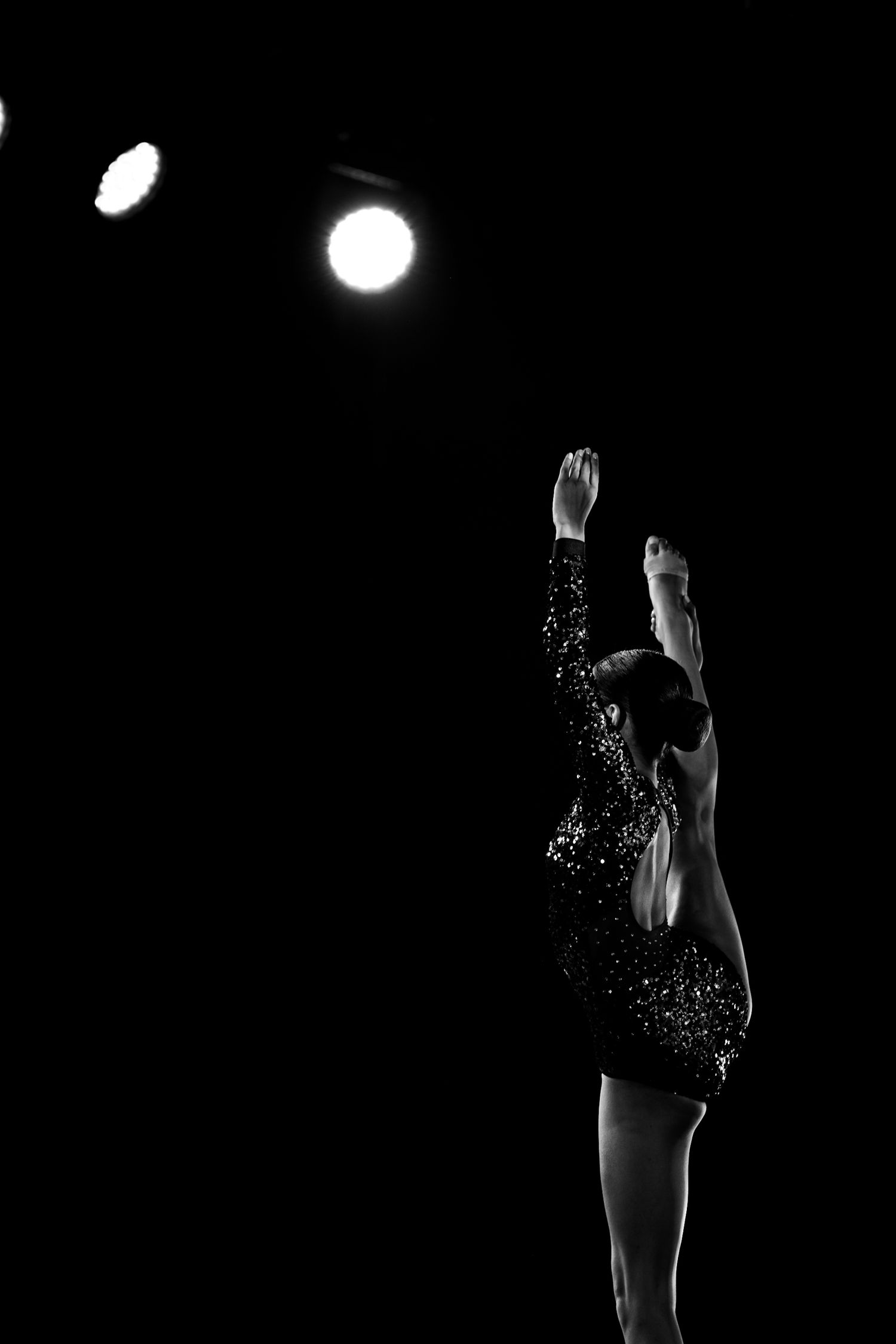 Black and white photo of a gymnast in a sparkling leotard reaching upward under bright stage spotlights.
