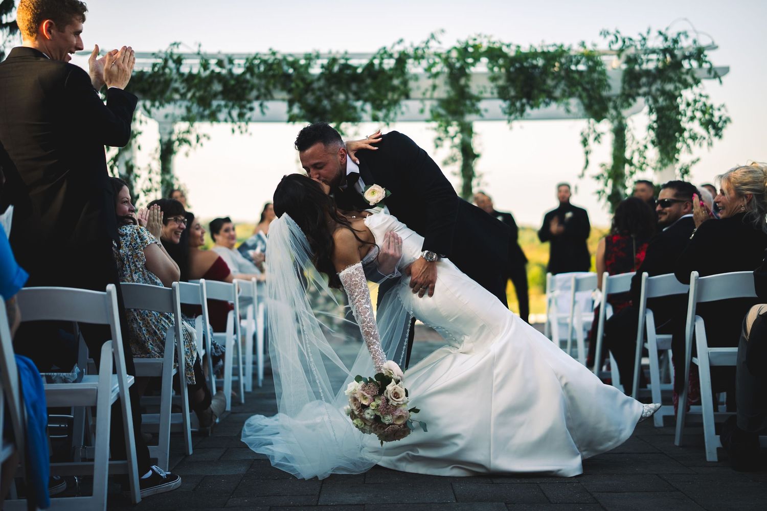 Groom dips bride for dramatic kiss at wedding ceremony altar with greenery arch and seated guests.