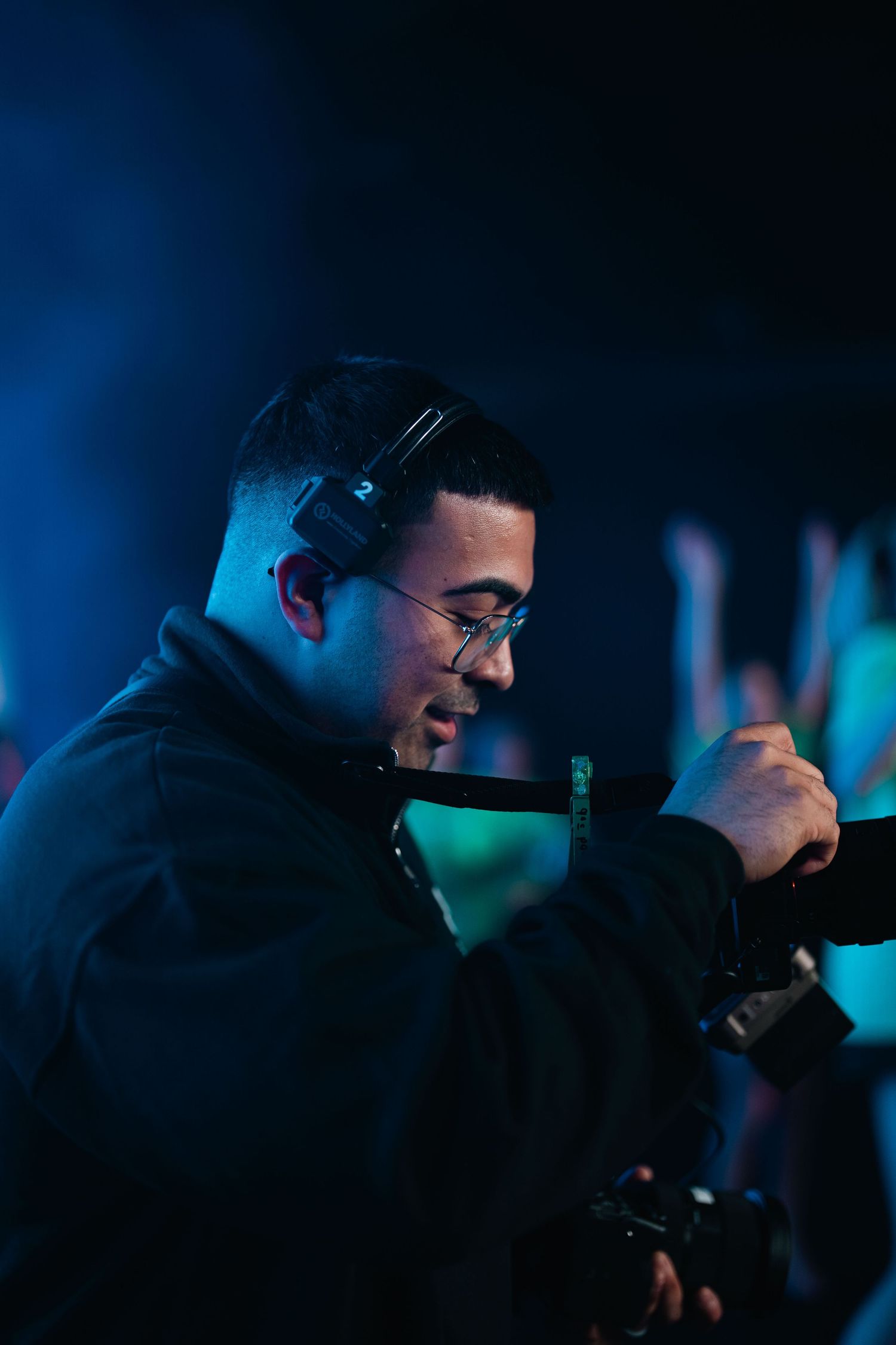 Male videographer with headphones and camera works at a vibrant event with blue neon lighting.