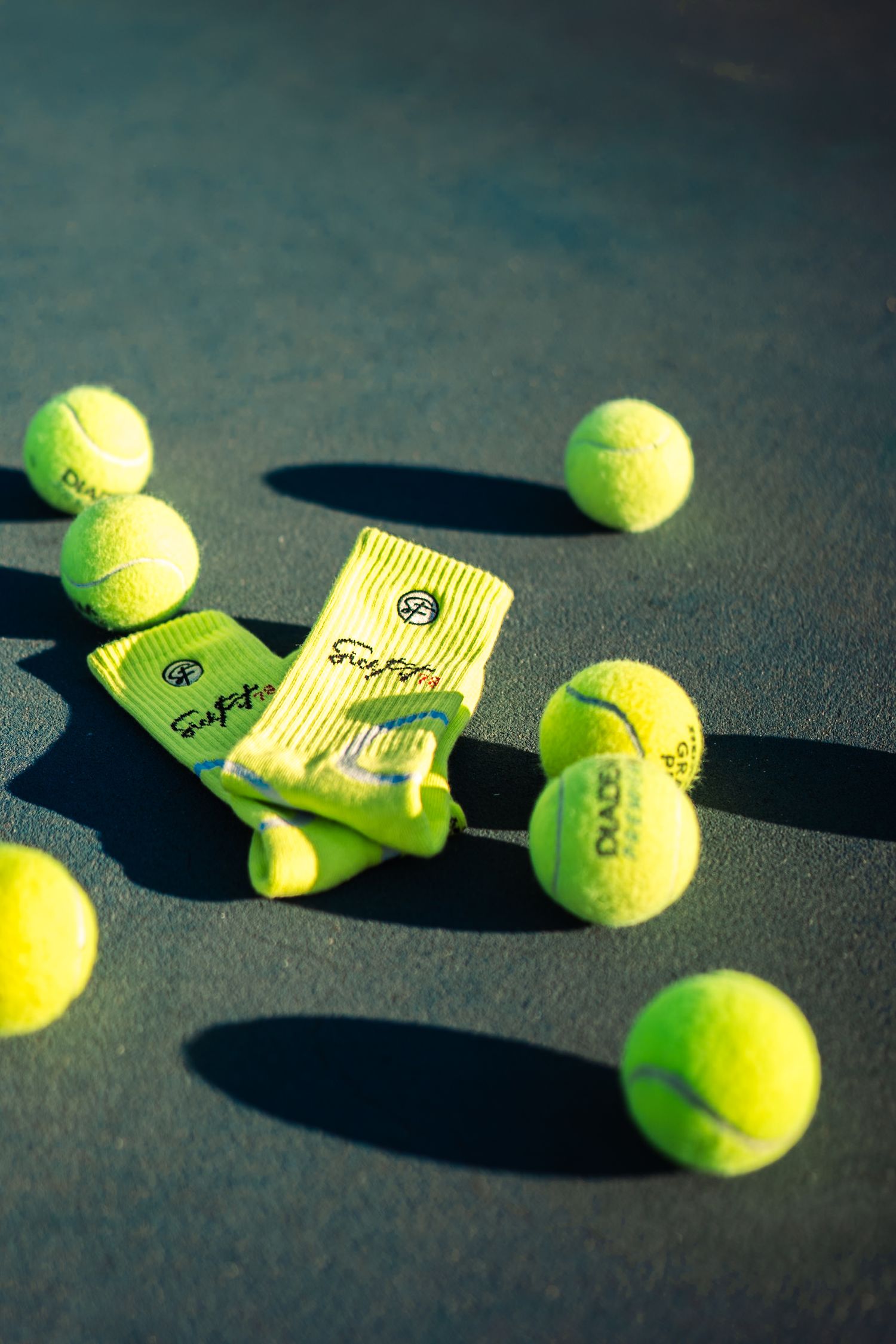 Neon yellow tennis socks placed among scattered tennis balls on a dark court surface in bright sunlight.