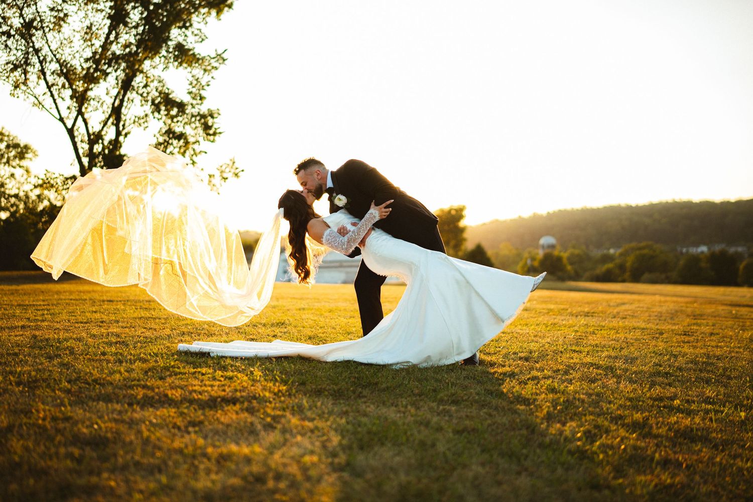 Bride and groom share romantic dip kiss at golden sunset on lush green lawn, veil flowing dramatically in the breeze.