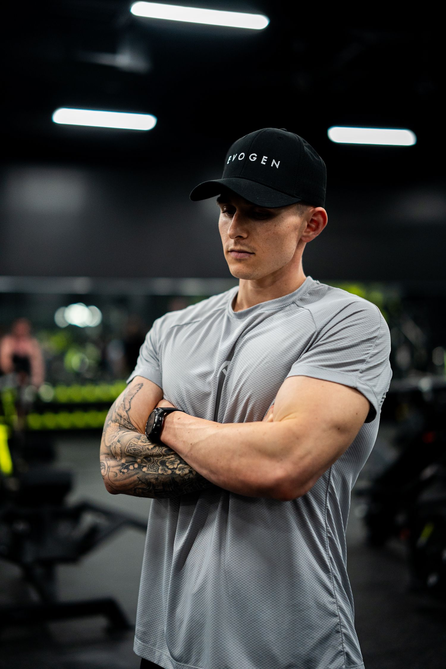 Muscular man in gray t-shirt and black cap stands with arms crossed in a modern gym, showcasing tattoos.