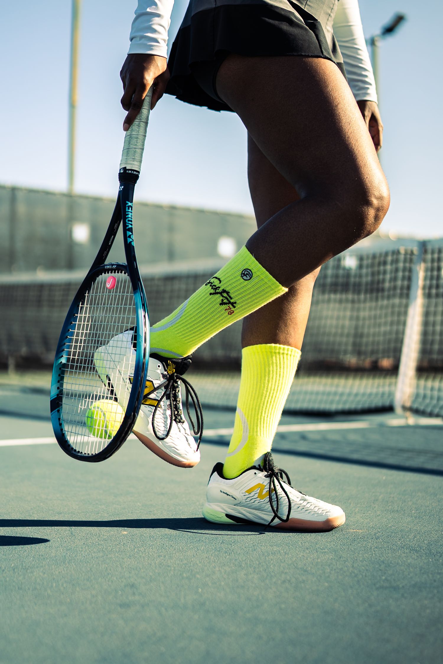 Tennis player in neon yellow socks and white sneakers holds racket on court with balls nearby on a sunny day.