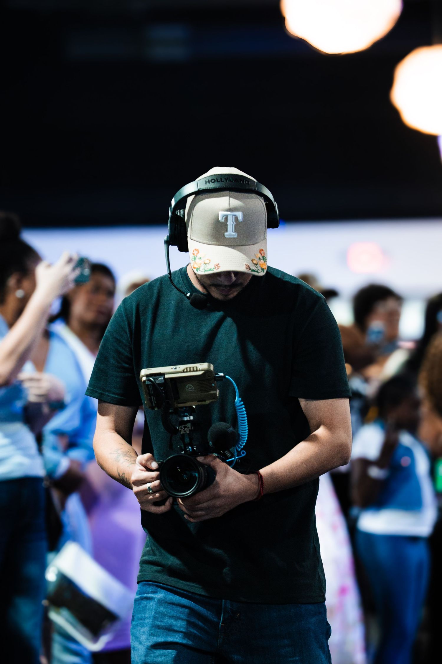 Cameraman holding professional camera equipment at a live event, wearing headset and snapback cap.