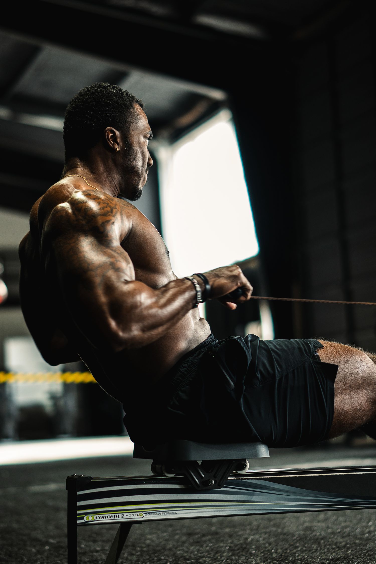 Muscular shirtless Black man performing cable row exercise on gym bench in dramatic lighting.