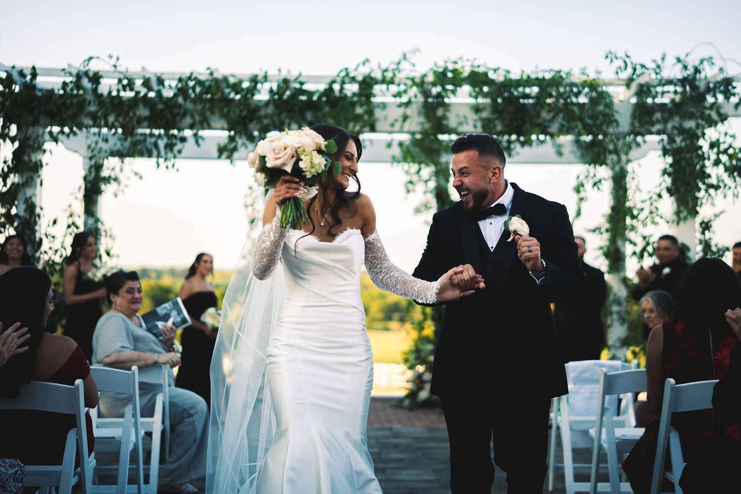Bride and groom walk down the aisle under a floral arch at an outdoor wedding ceremony.