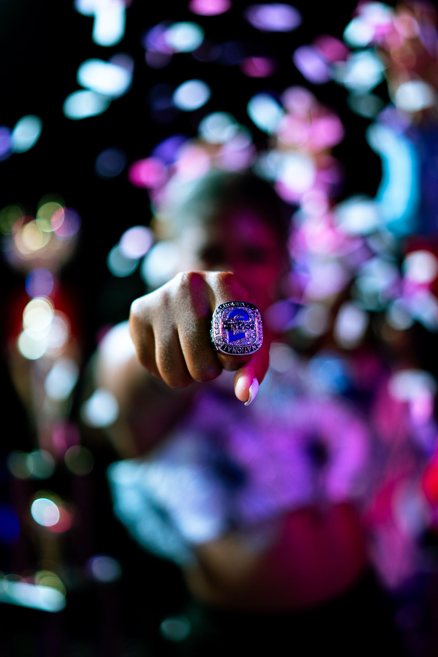 A hand wearing a large championship ring is held up toward the camera against a colorful bokeh background.