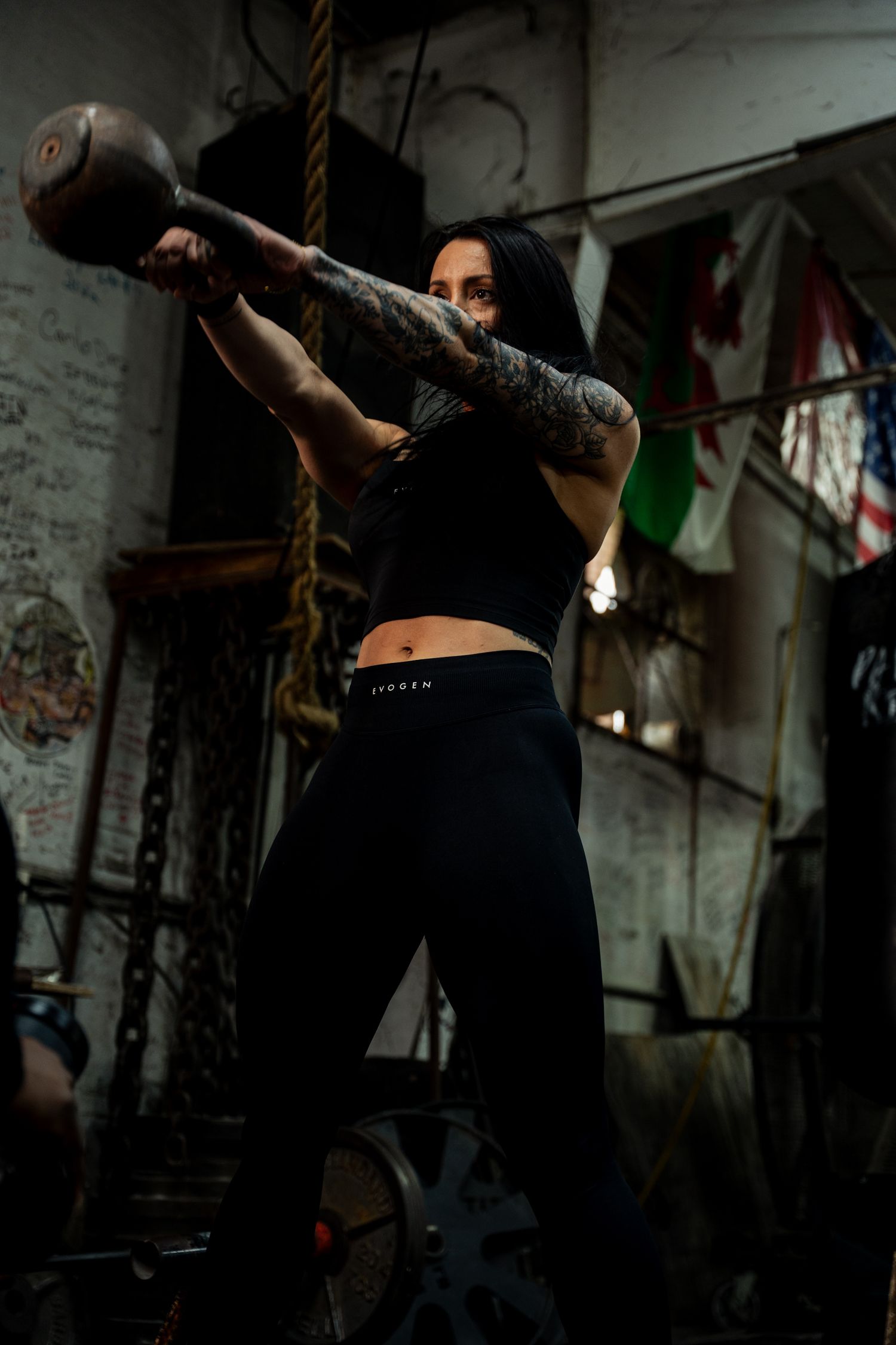 Tattooed woman in black Evogen crop top swinging kettlebell in dark industrial gym setting.