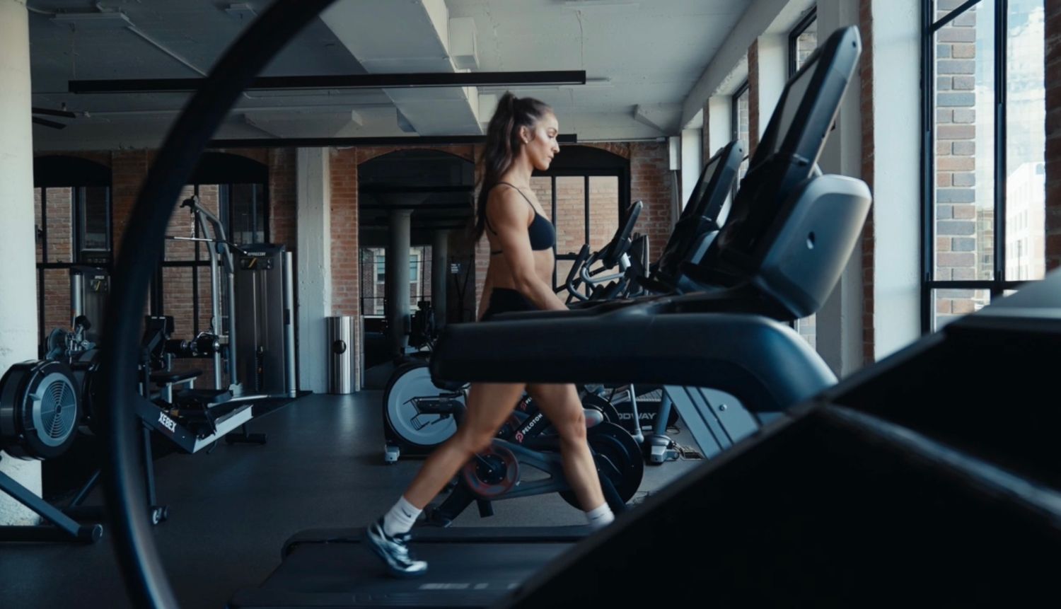Woman running on treadmill in modern gym with equipment and natural light from windows.