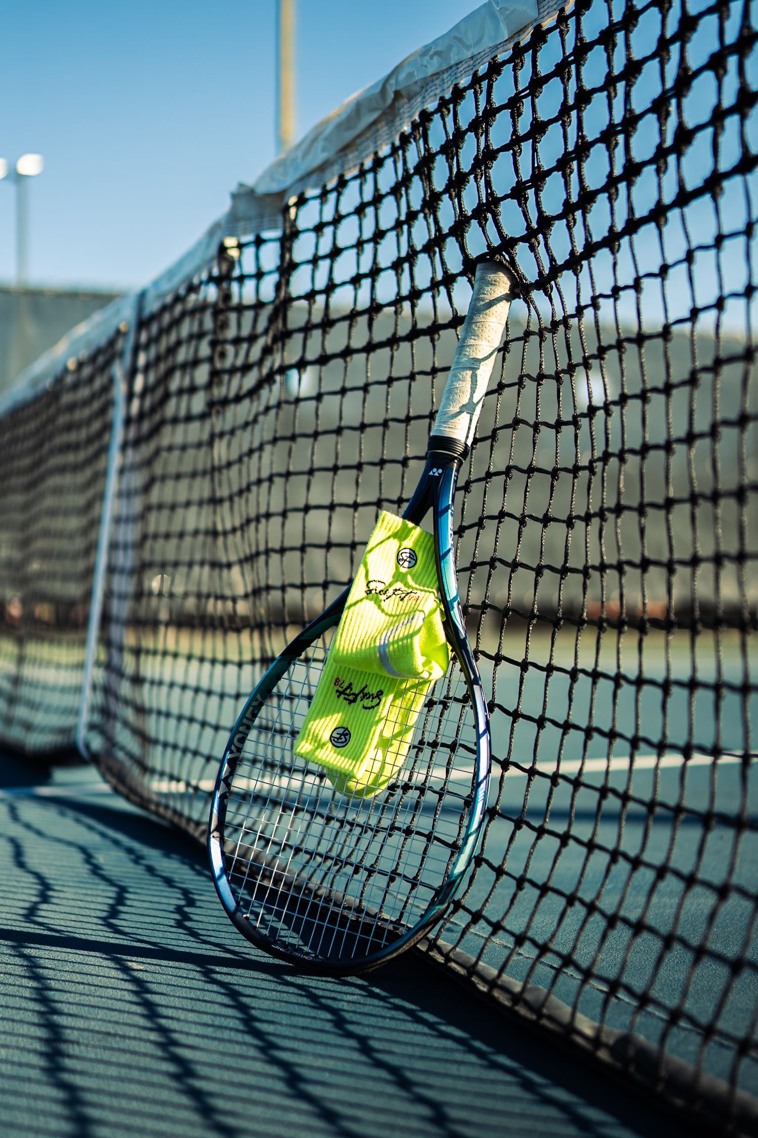 Tennis racket with yellow balls leaning against a black net on an outdoor court in bright sunlight.