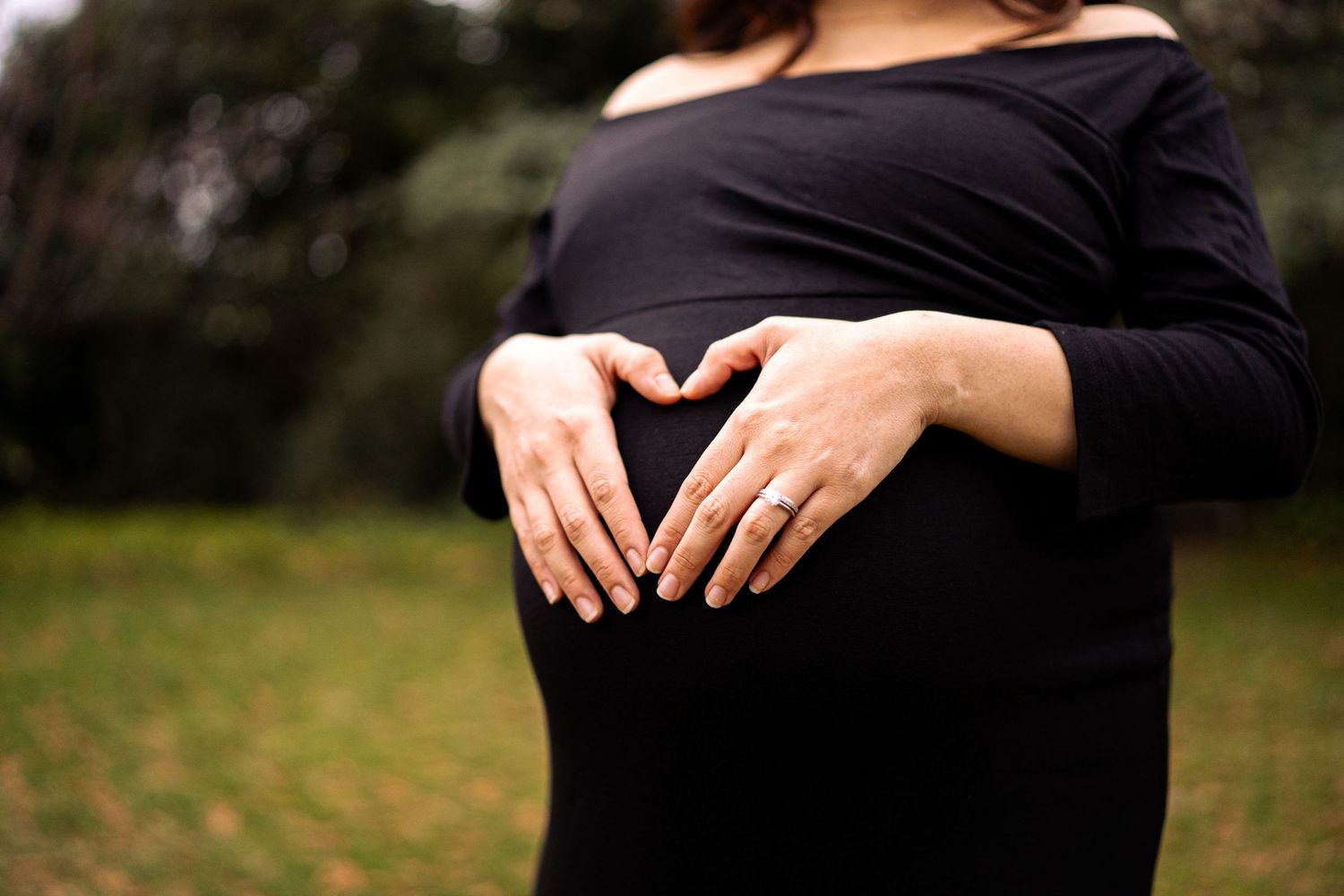 Pregnant woman in black dress forming heart shape with hands on baby bump outdoors.