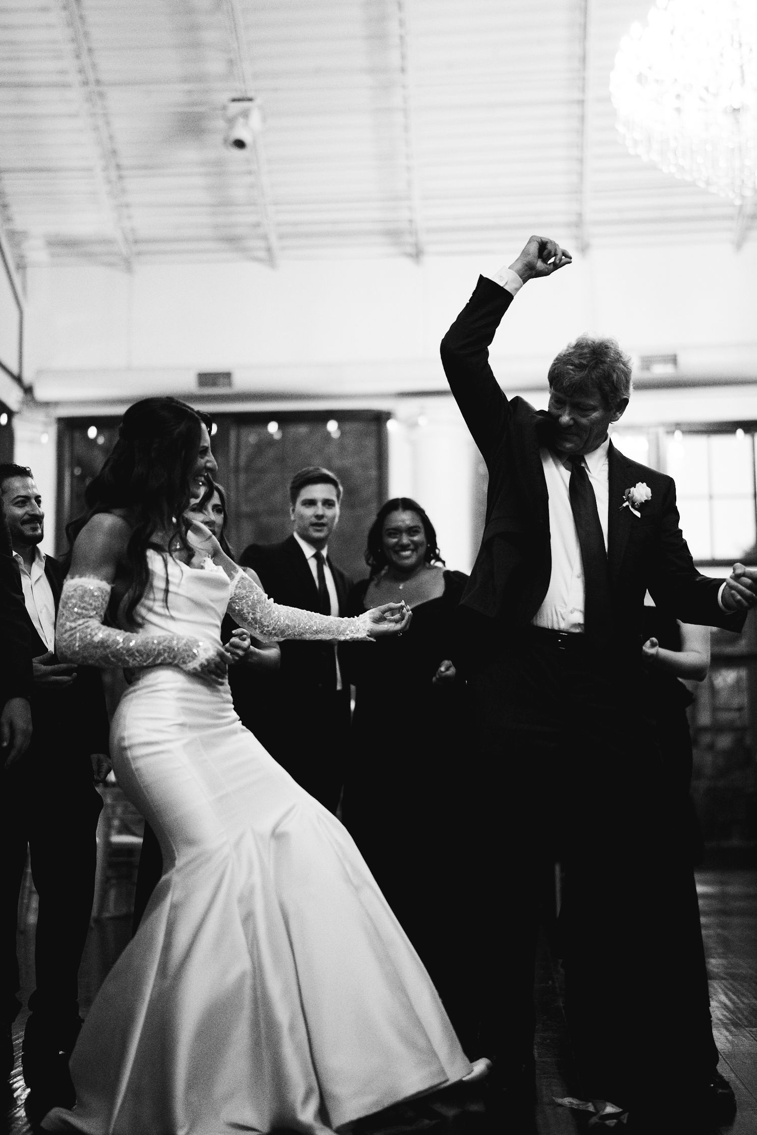 Black and white photo of bride and groom dancing energetically with guests watching at reception.