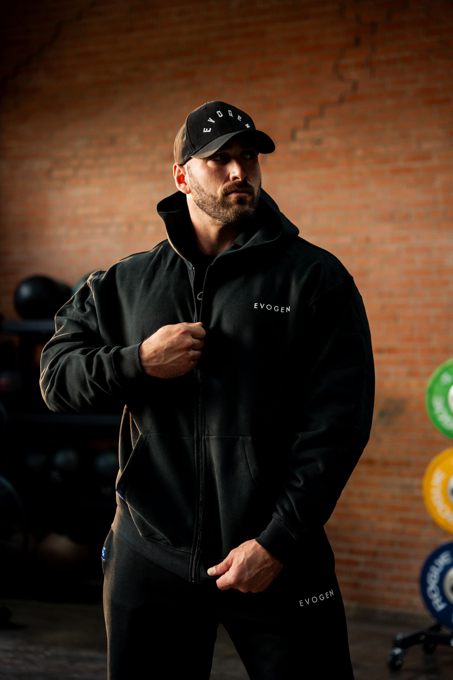 Muscular man wearing black Evogen zip-up hoodie and cap posing confidently in gym setting.