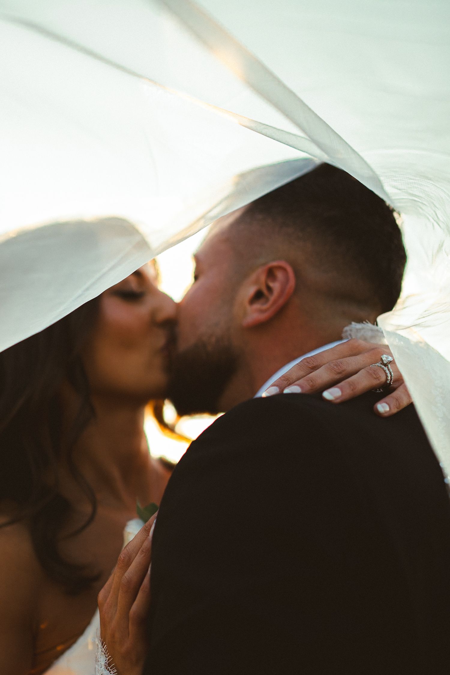 Bride and groom share passionate kiss under wedding veil in glowing backlit sunset photography.