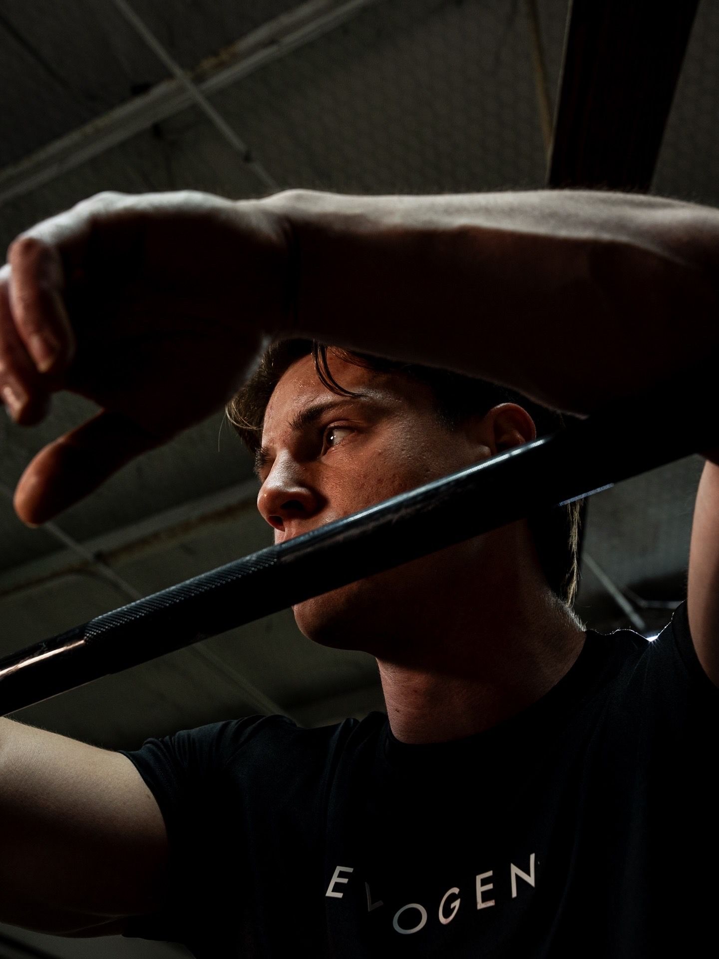 Athletic man in black Evogen t-shirt gripping barbell during intense weightlifting session in gym.
