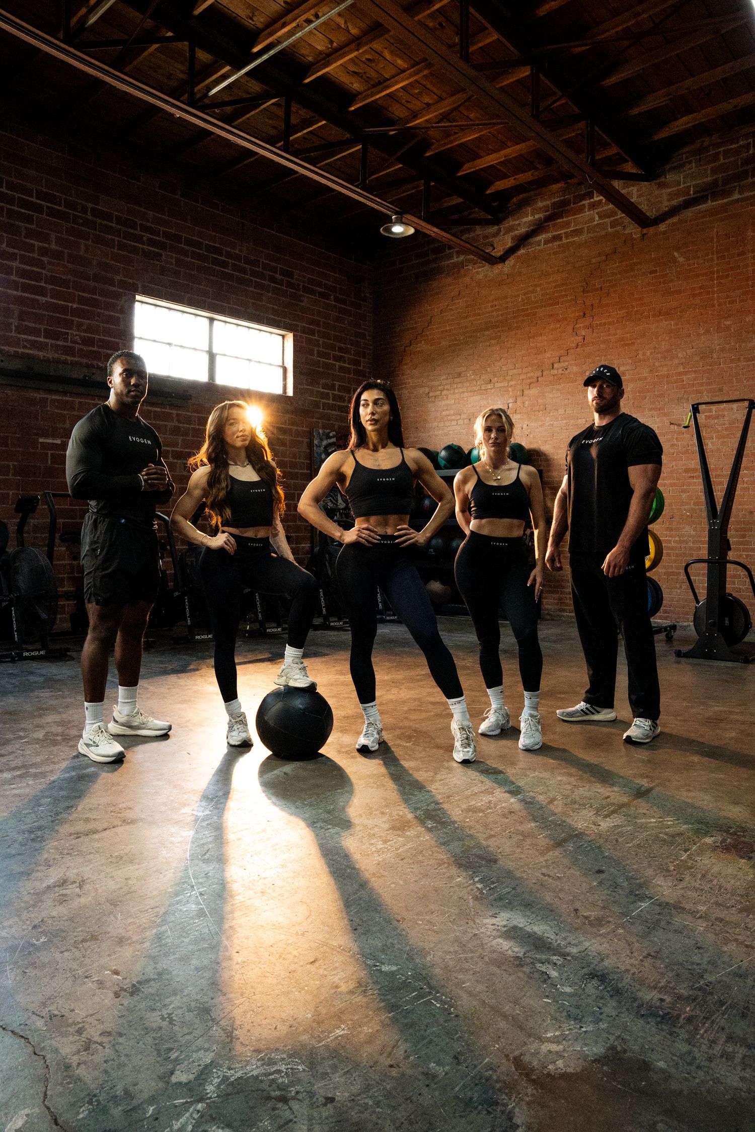 Group of five fit athletes in black workout gear posing in industrial brick gym with medicine ball.