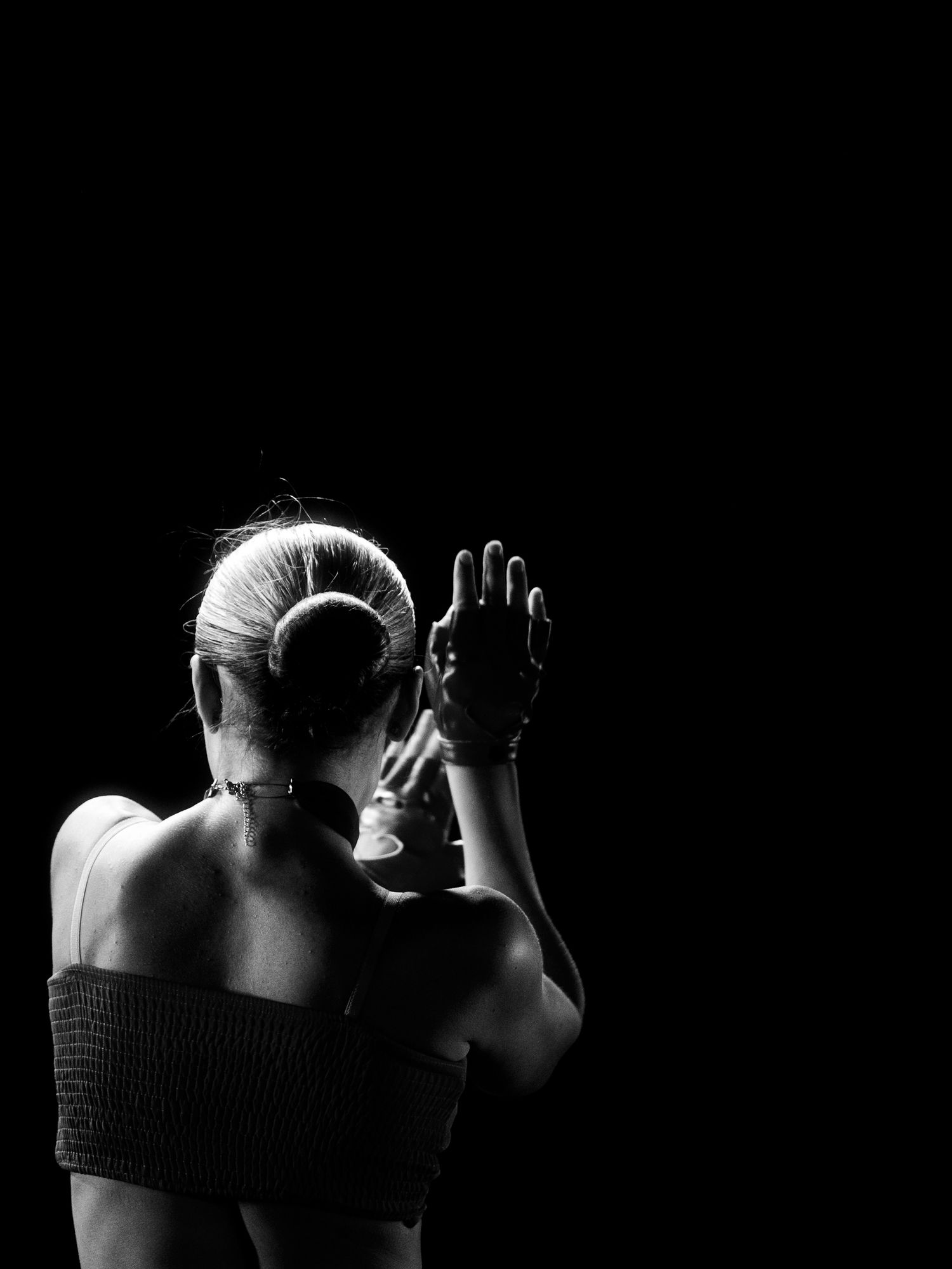 Black and white photo of a woman with a bun hairstyle, seen from behind, raising her hand against a dark background.