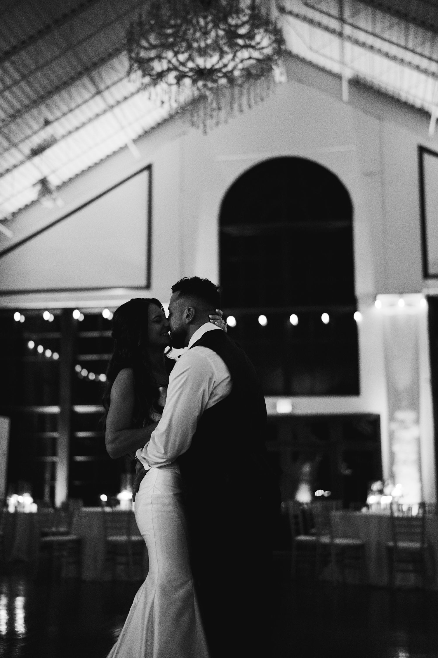 Black and white photo of bride and groom sharing a kiss during their first dance indoors.