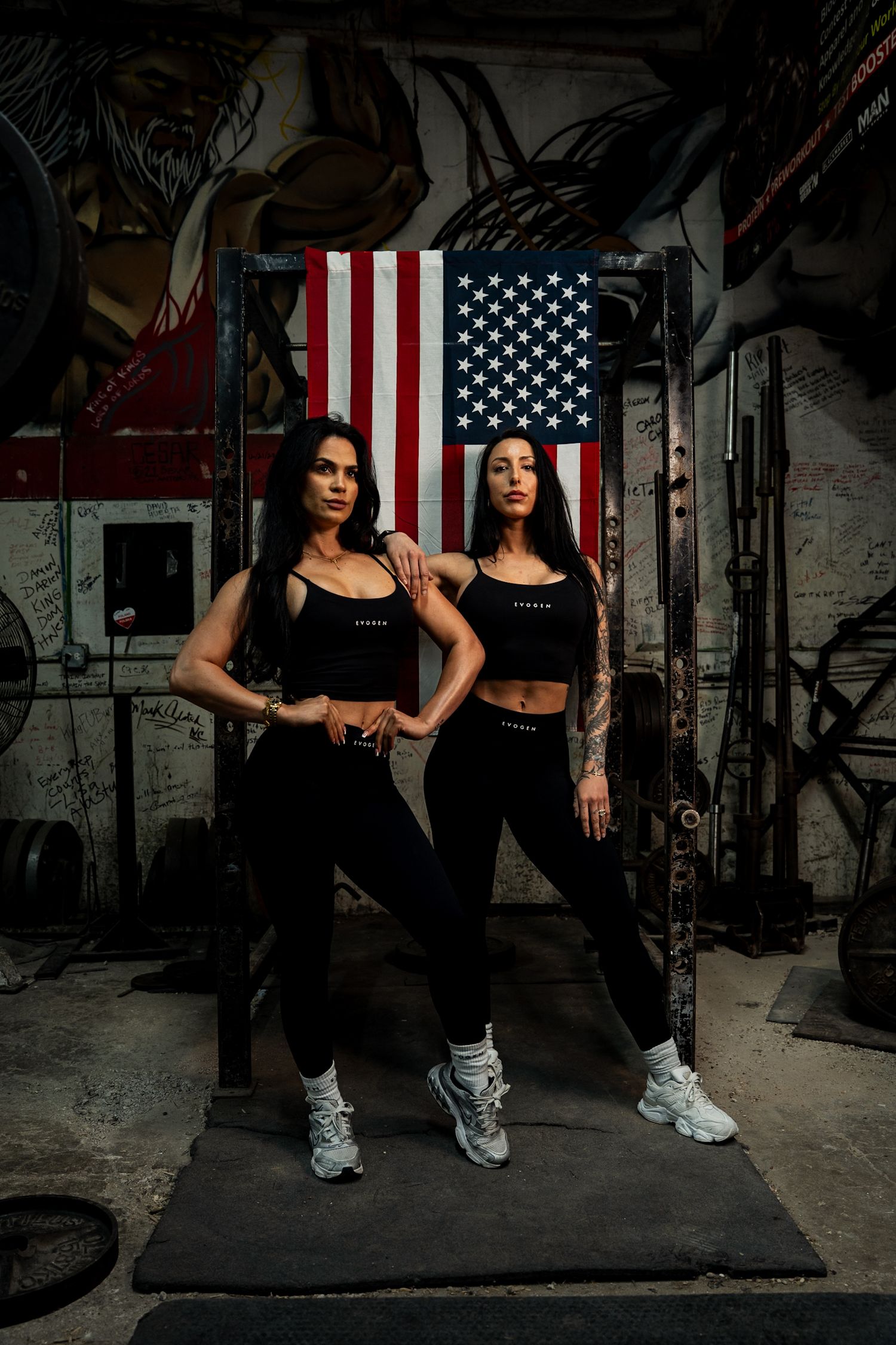 Two fit women in black Evogen activewear pose confidently in a gym with an American flag backdrop.