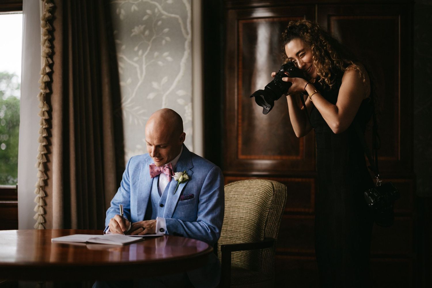 Photographer captures candid moment of someone in blue suit writing at wooden desk by window with elegant curtains.