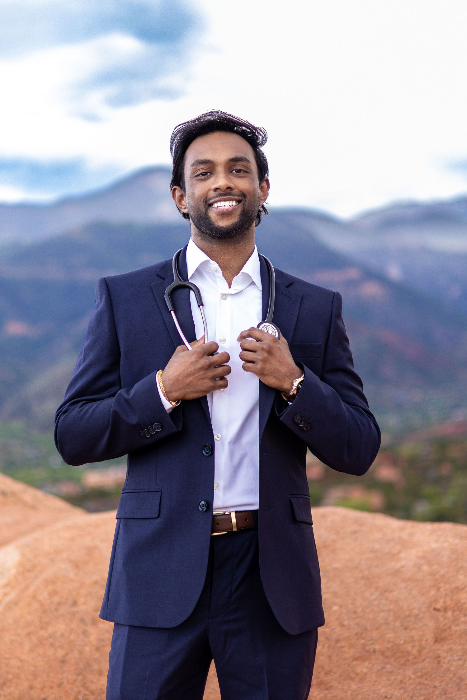 Smiling Indian male doctor in navy suit with stethoscope poses outdoors against scenic mountain backdrop.