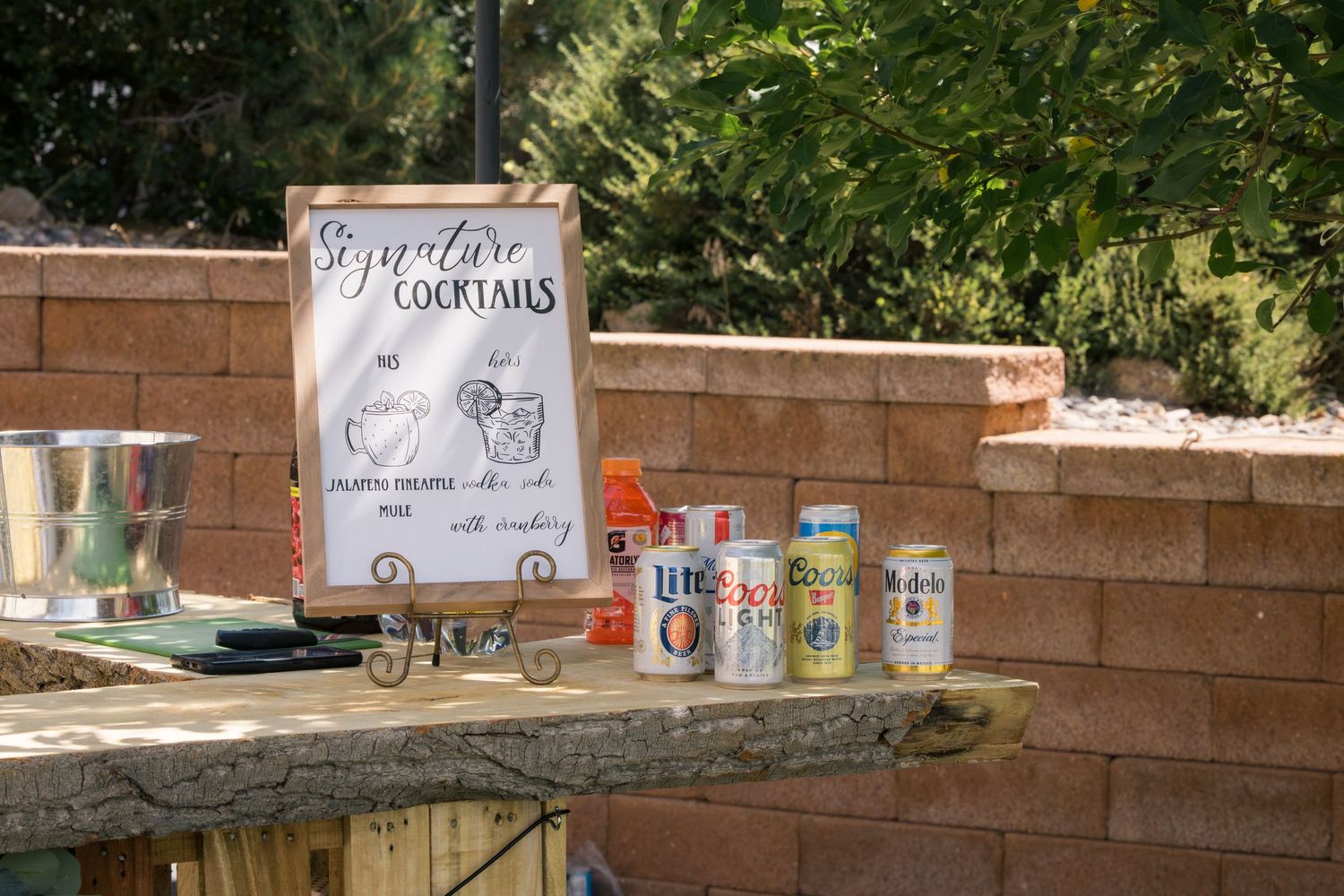 Outdoor signature cocktails sign displayed on a wooden table with beer cans beside a brick wall at a party.