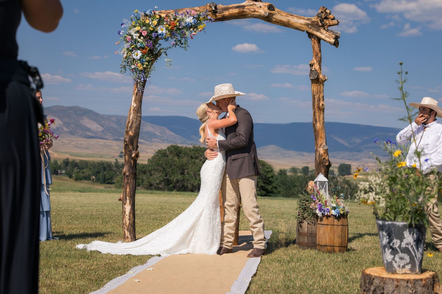 Couple shares first kiss under rustic wooden arch at outdoor wedding ceremony with mountain views.